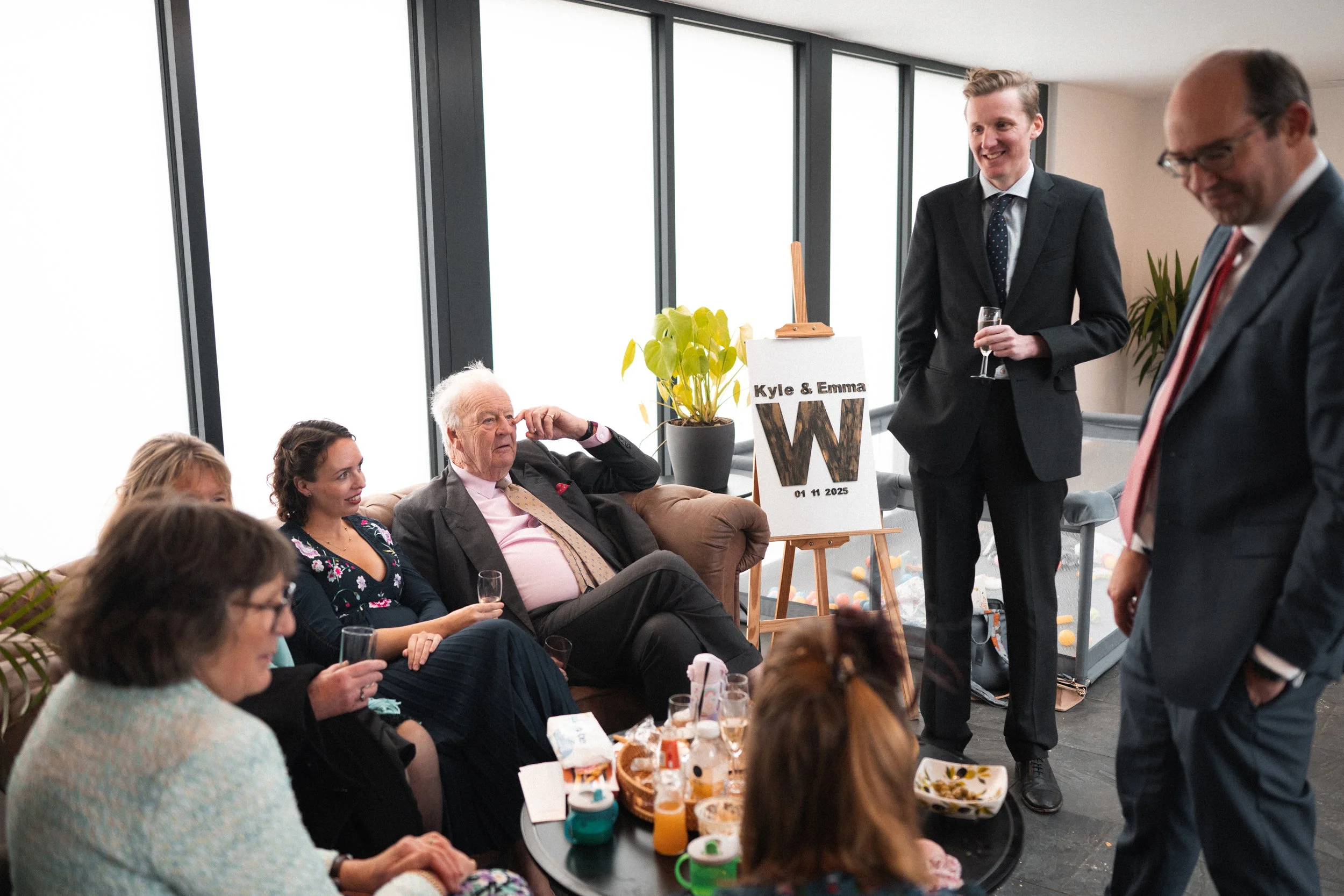 People enjoying a celebration in an indoor lounge next to large windows. A man in a suit is speaking, holding a glass of champagne. There is a sign with the names "Kyle & Emma" and the date 01/11/2025. Several guests are sitting and standing, holding