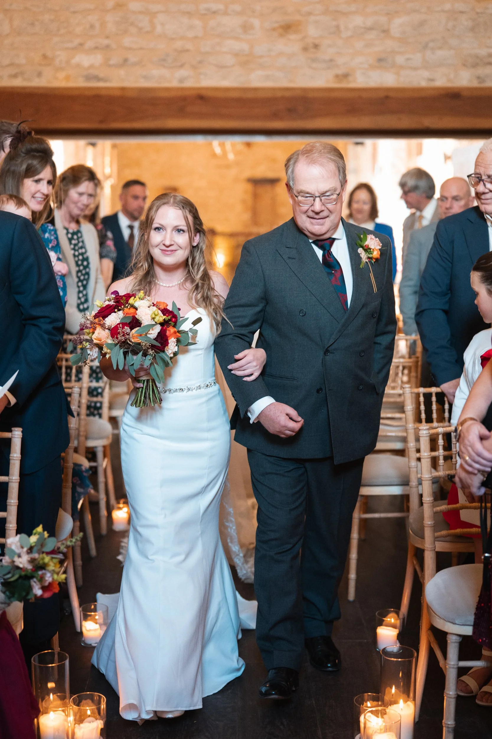 A wedding ceremony with a bride in a white gown carrying a bouquet, walking down the aisle with an older man in a dark suit, surrounded by seated guests and lit candles.