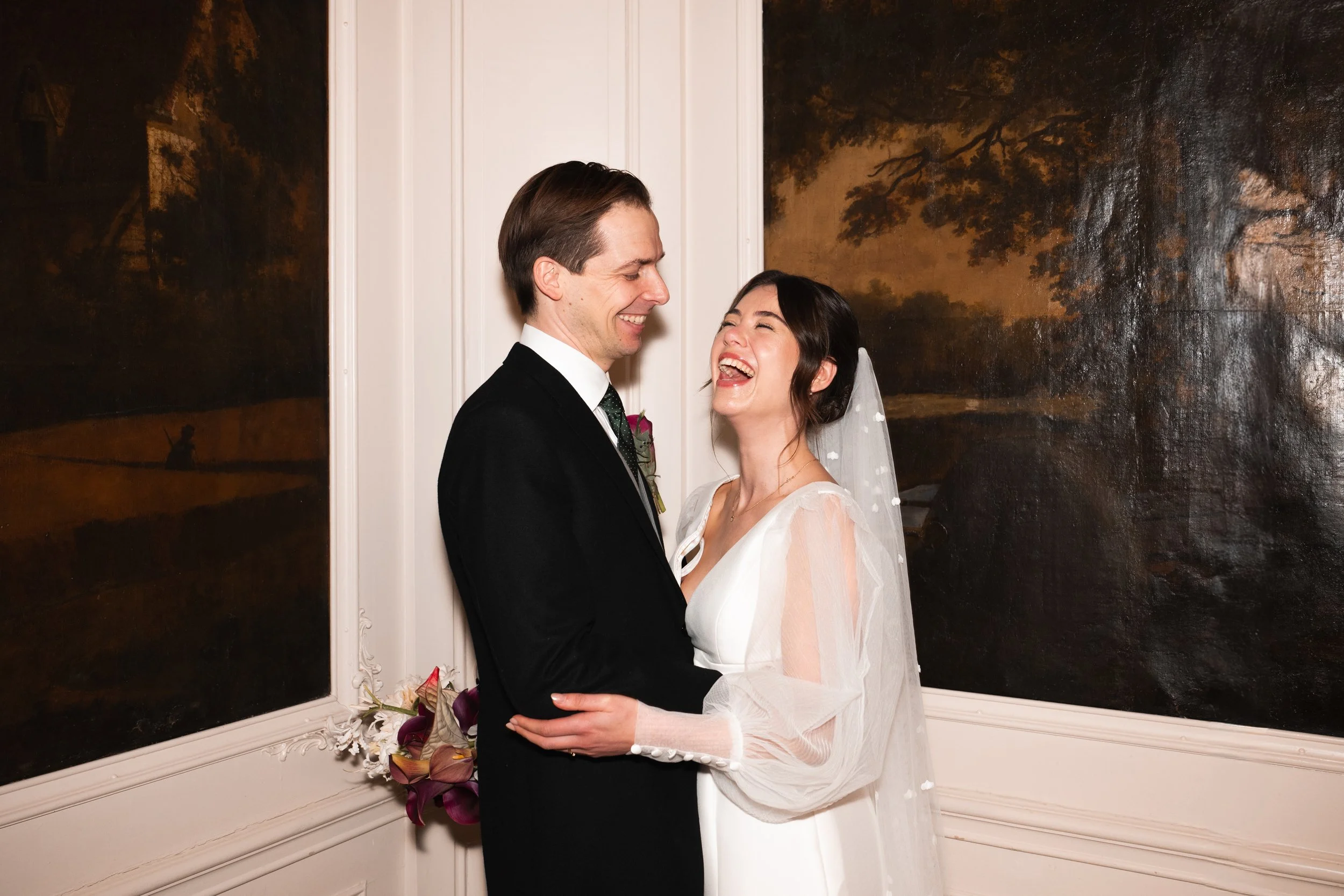 A newlywed couple sharing a joyful moment indoors, standing close to each other in front of dark landscape paintings, with the bride in a white wedding dress and veil, and the groom in a black suit and tie.