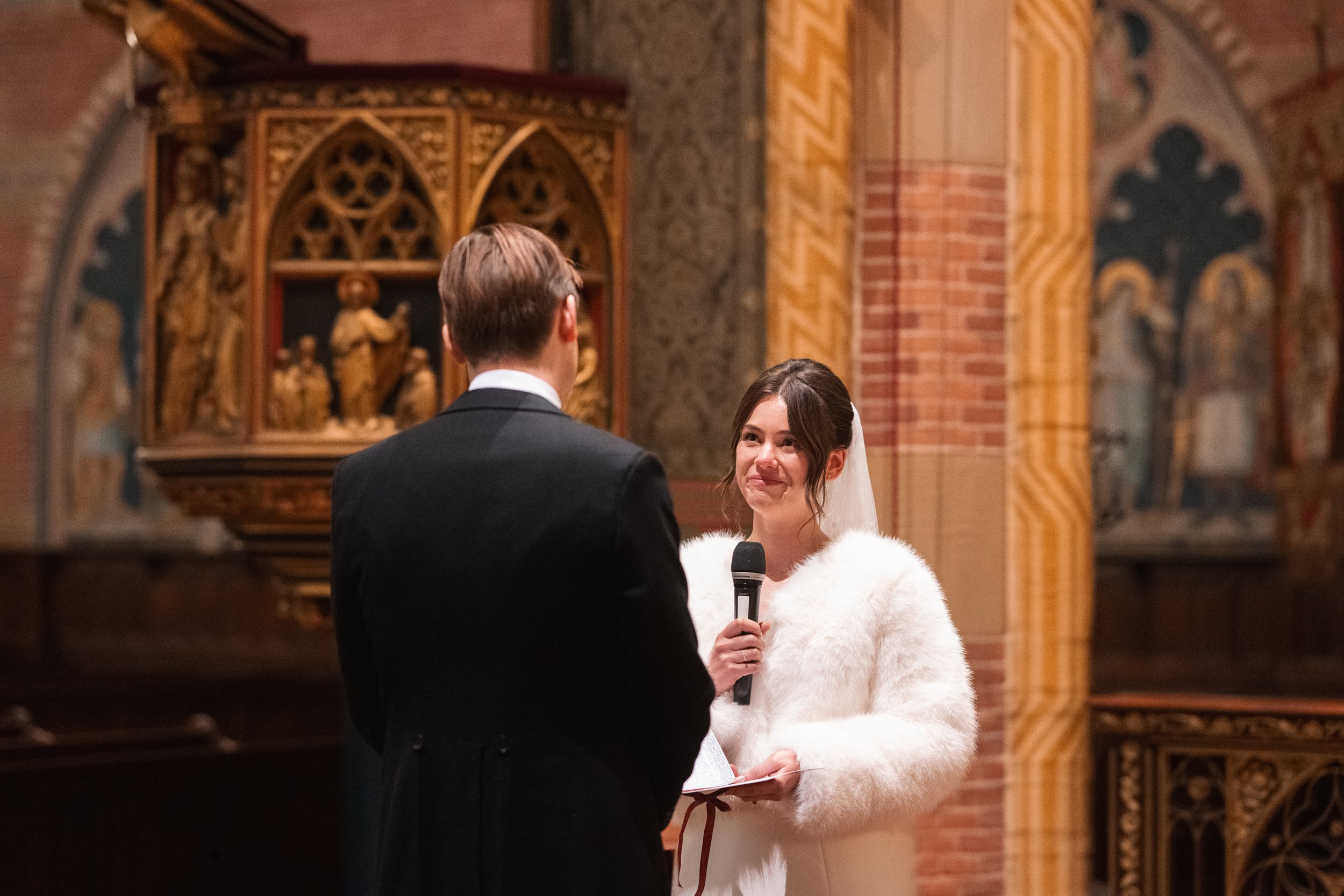 A woman in a white dress and faux fur coat holding a microphone appears to be officiating a wedding ceremony in a church, facing a man in a black suit with his back to the camera, with ornate religious artwork in the background.