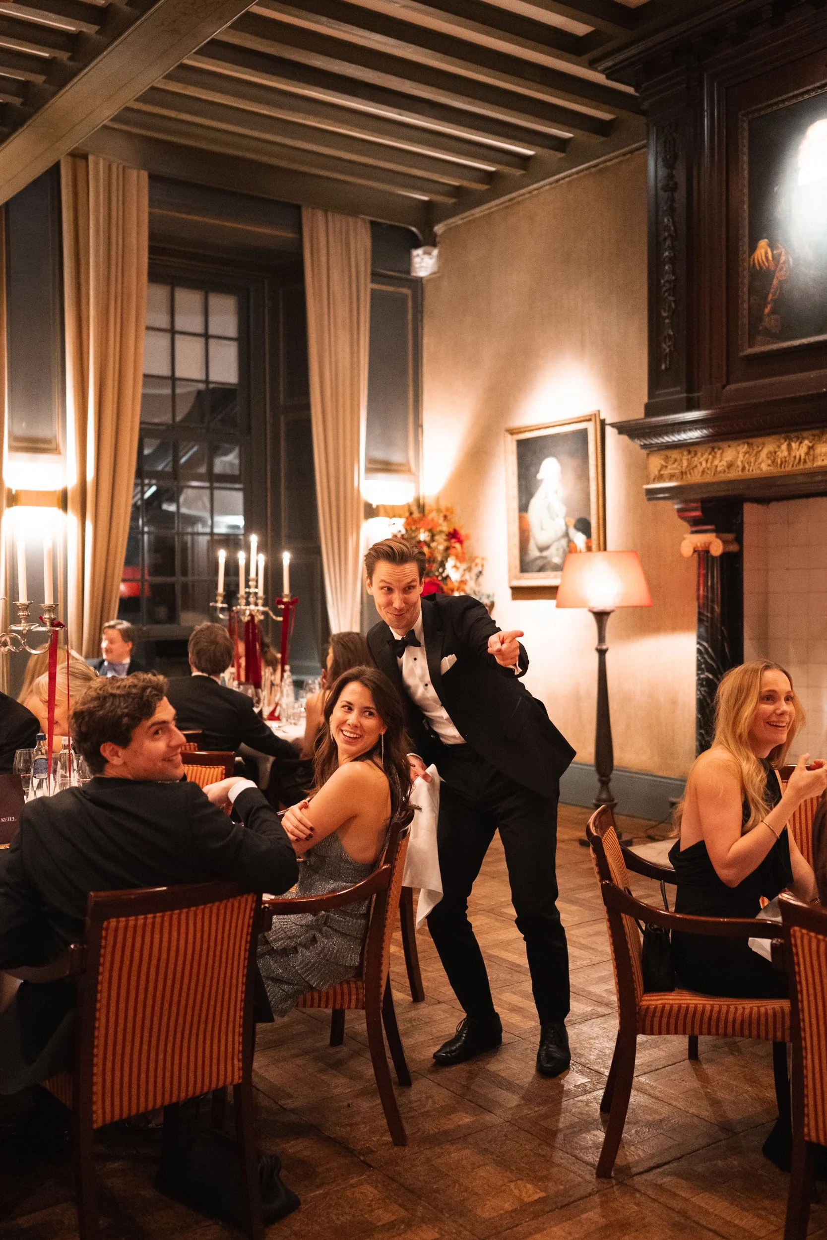 A waiter in a tuxedo points and poses playfully at a formal dinner party in an elegant, warmly lit room with classic artwork, tall candles, and draped curtains.