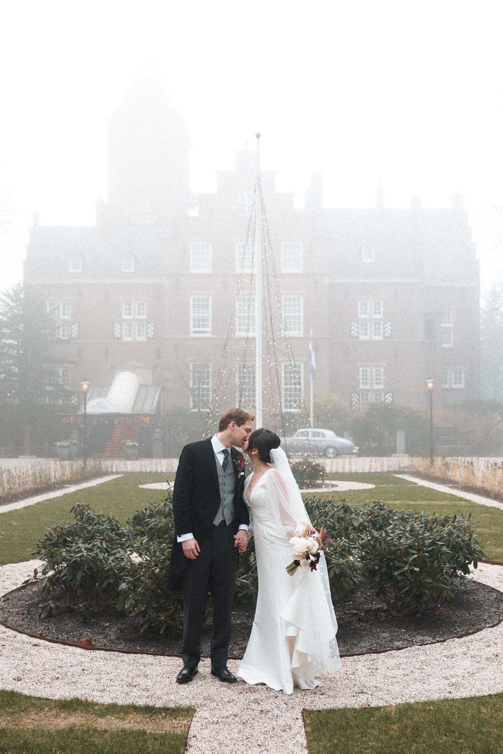 A bride and groom stand close together, holding hands and sharing a kiss outdoors on their wedding day. The bride is in a white wedding dress holding a bouquet, and the groom is in a dark suit with a gray vest and a boutonniere. Behind them is a smal