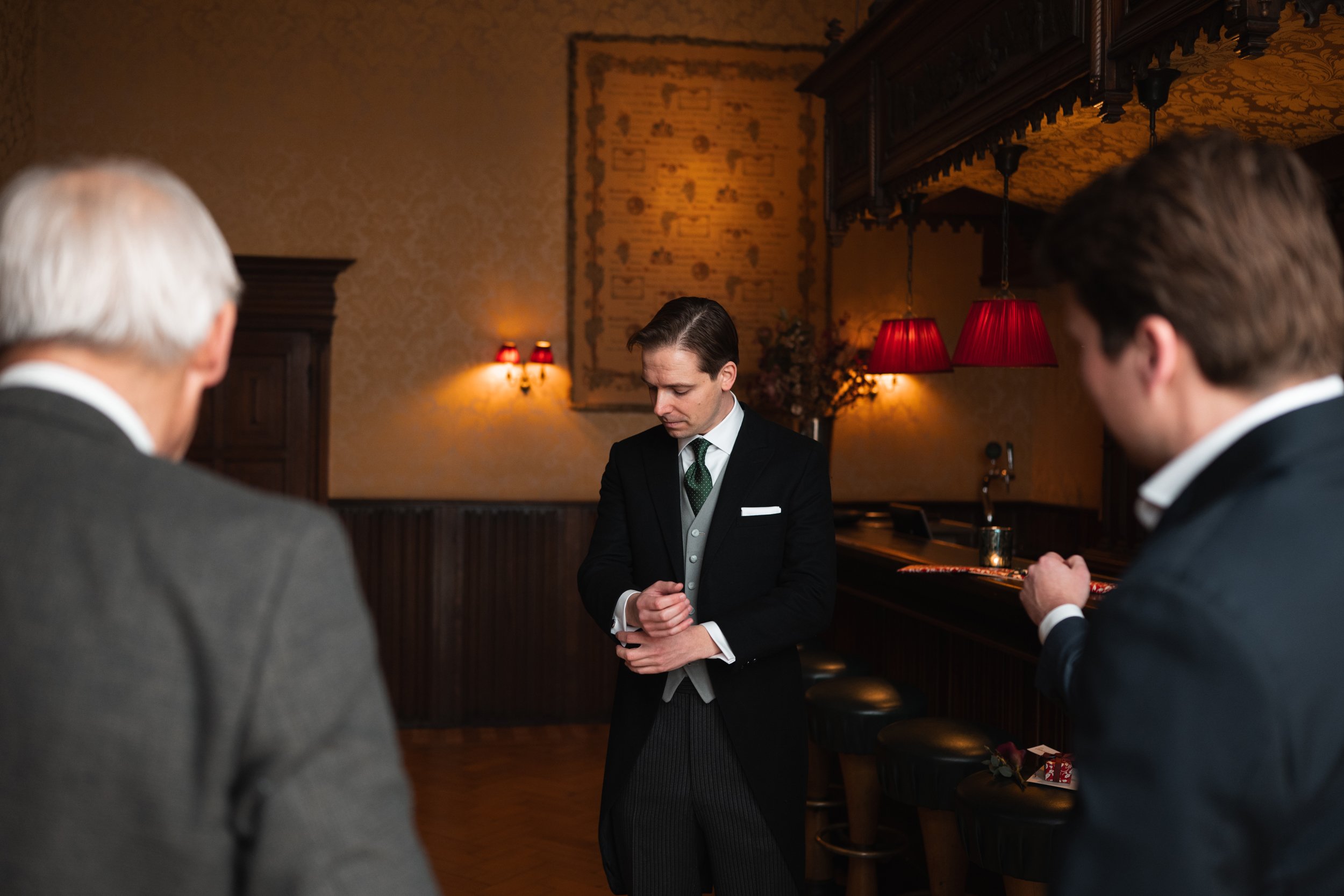 A man in a tuxedo bowing with hands clasped while two men in suits stand on either side watching, inside a warmly lit room with red lamps and vintage decor.
