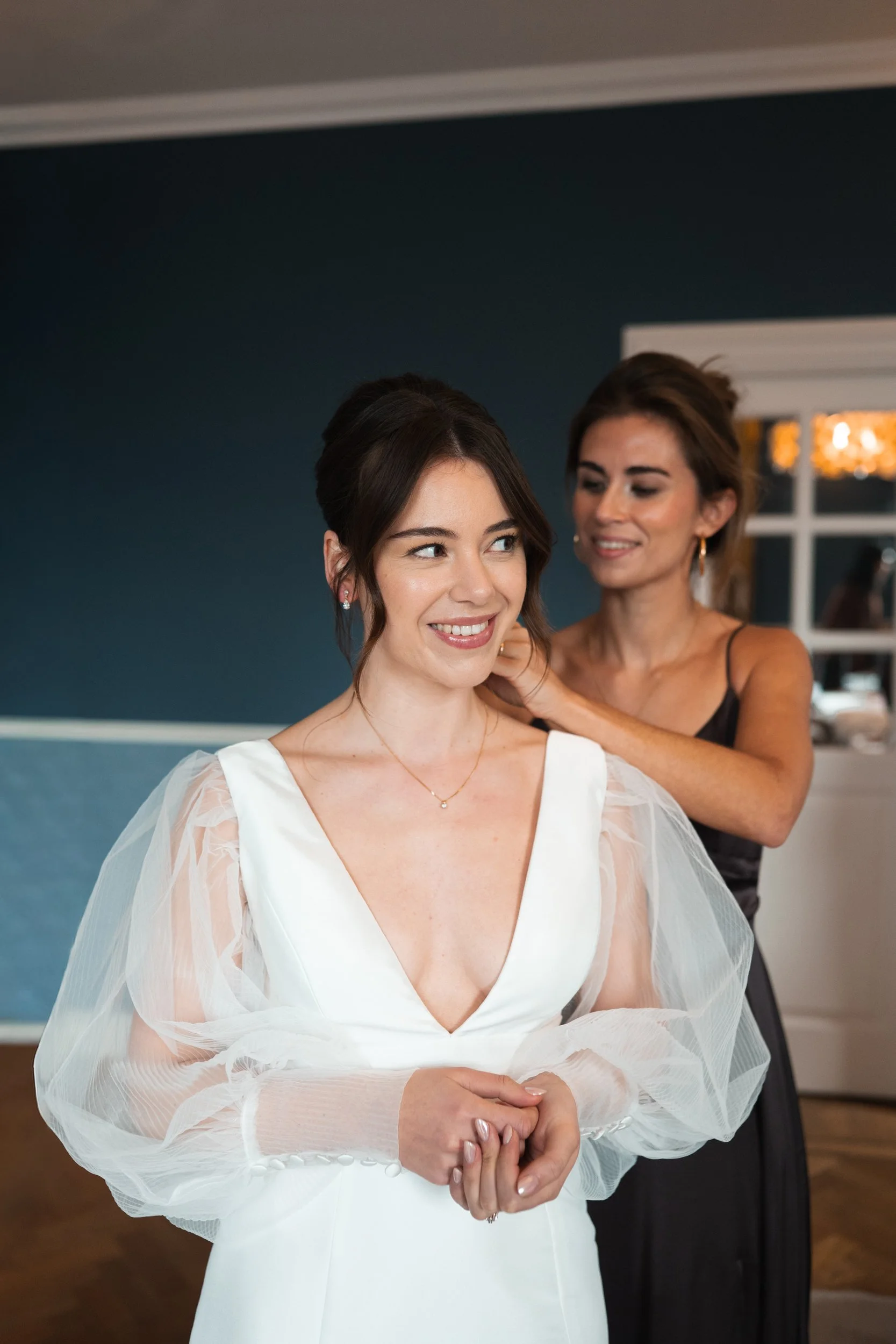 A bride in a white dress with sheer puffy sleeves gets ready as another woman helps adjust her hair, both smiling in a room with blue walls.