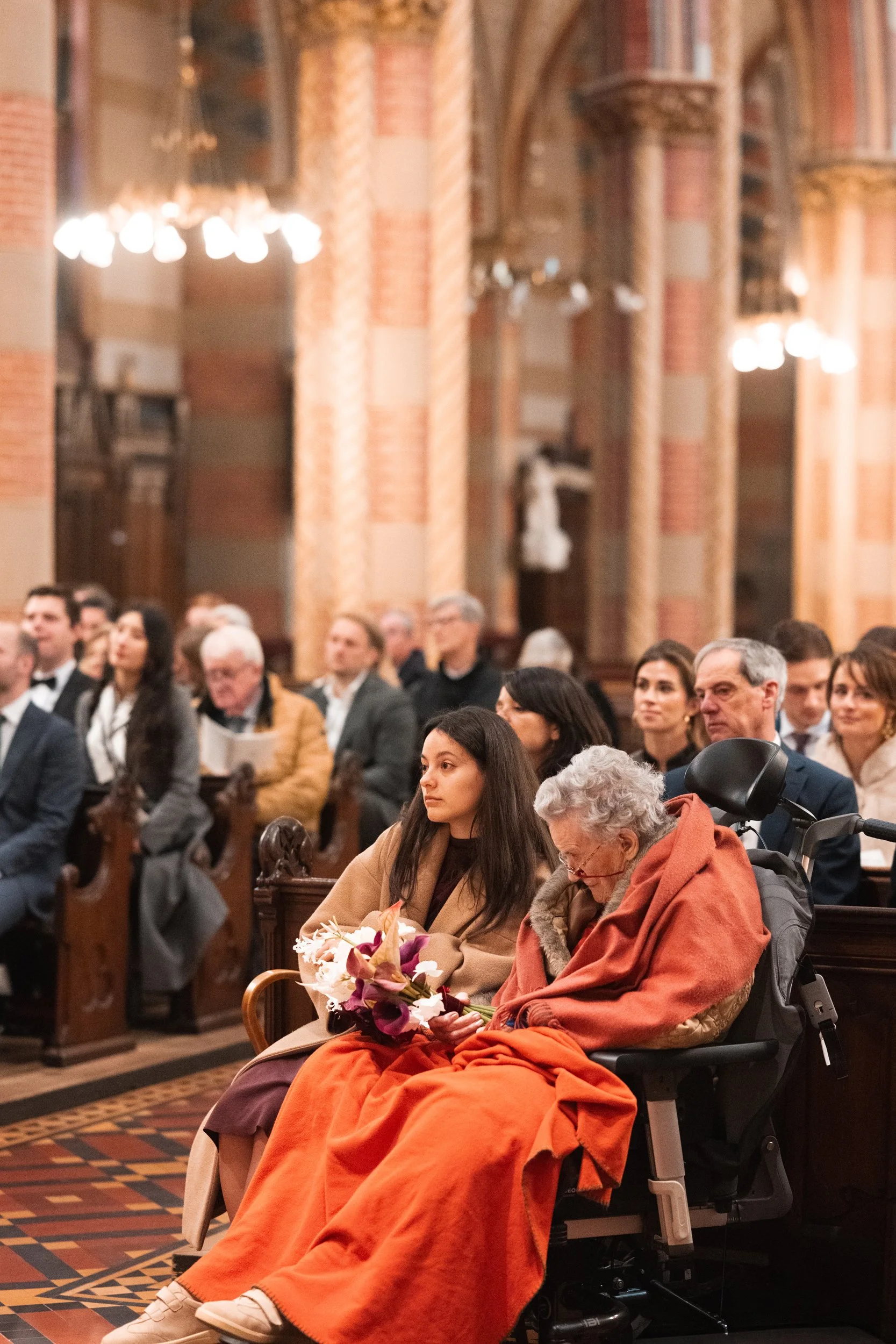 People seated inside a church, attending a ceremony or service. An older woman in a wheelchair is holding a bouquet of flowers, with a young woman sitting beside her.