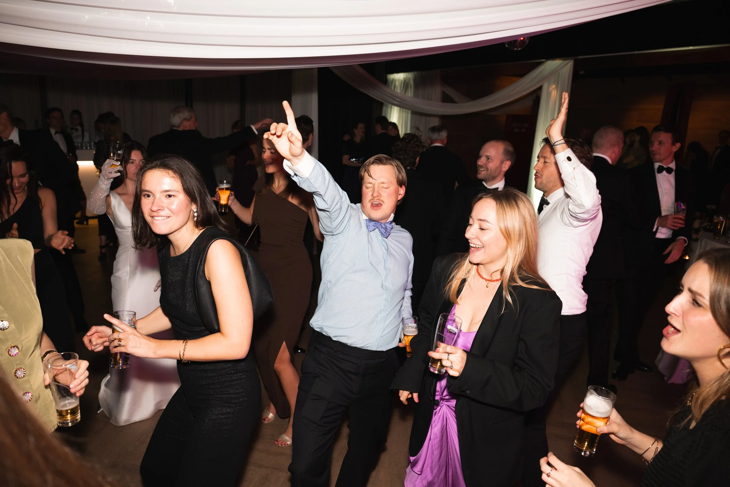People dancing and enjoying drinks at a celebration party in an indoor venue with decorated ceiling and curtains.