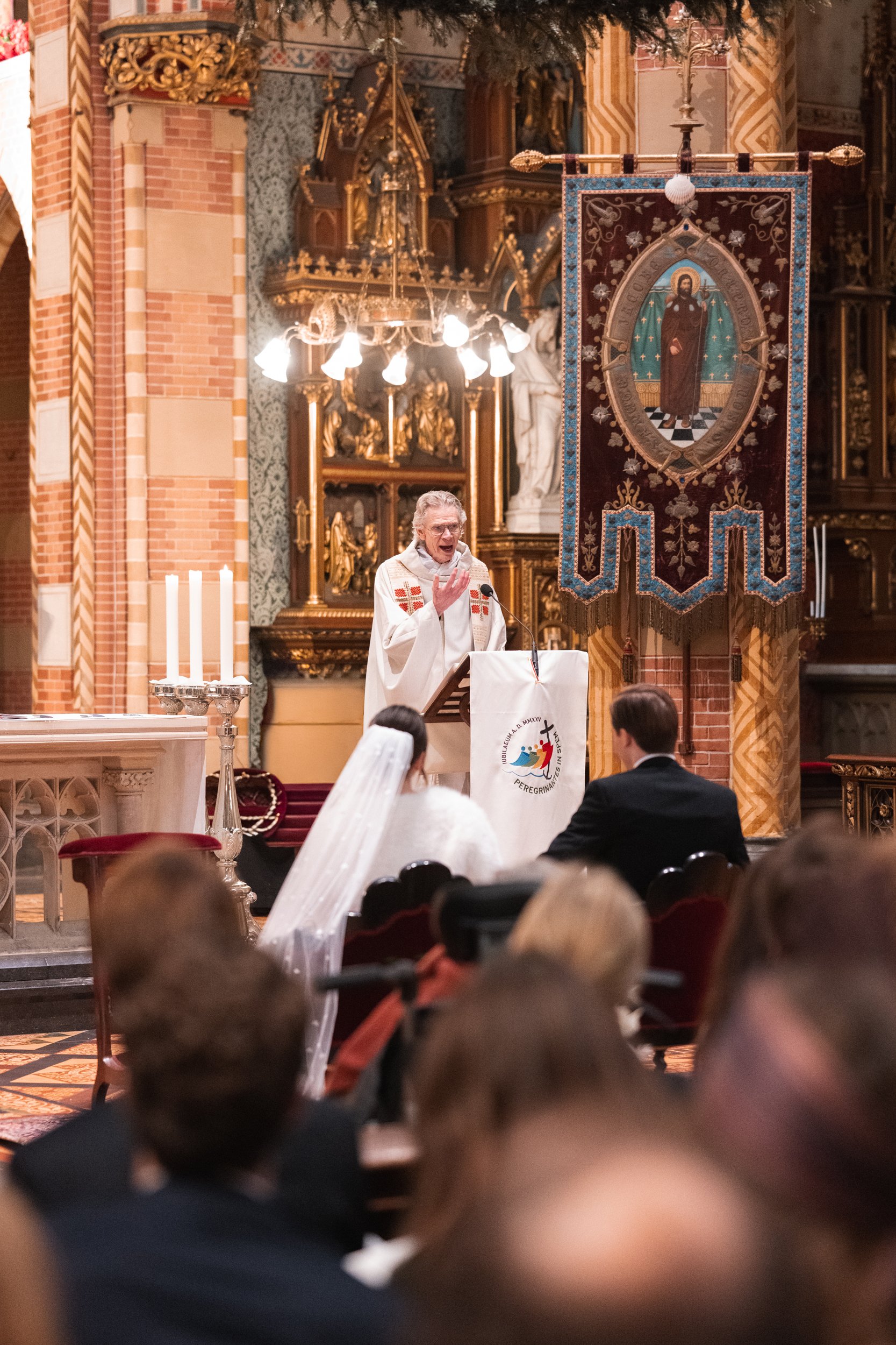 A priest giving a sermon at an altar in a church with gold accents, stained glass, and a large banner depicting a saint. Several people, including a bride in a white dress and veil, and a groom in a black suit, are seated and listening.