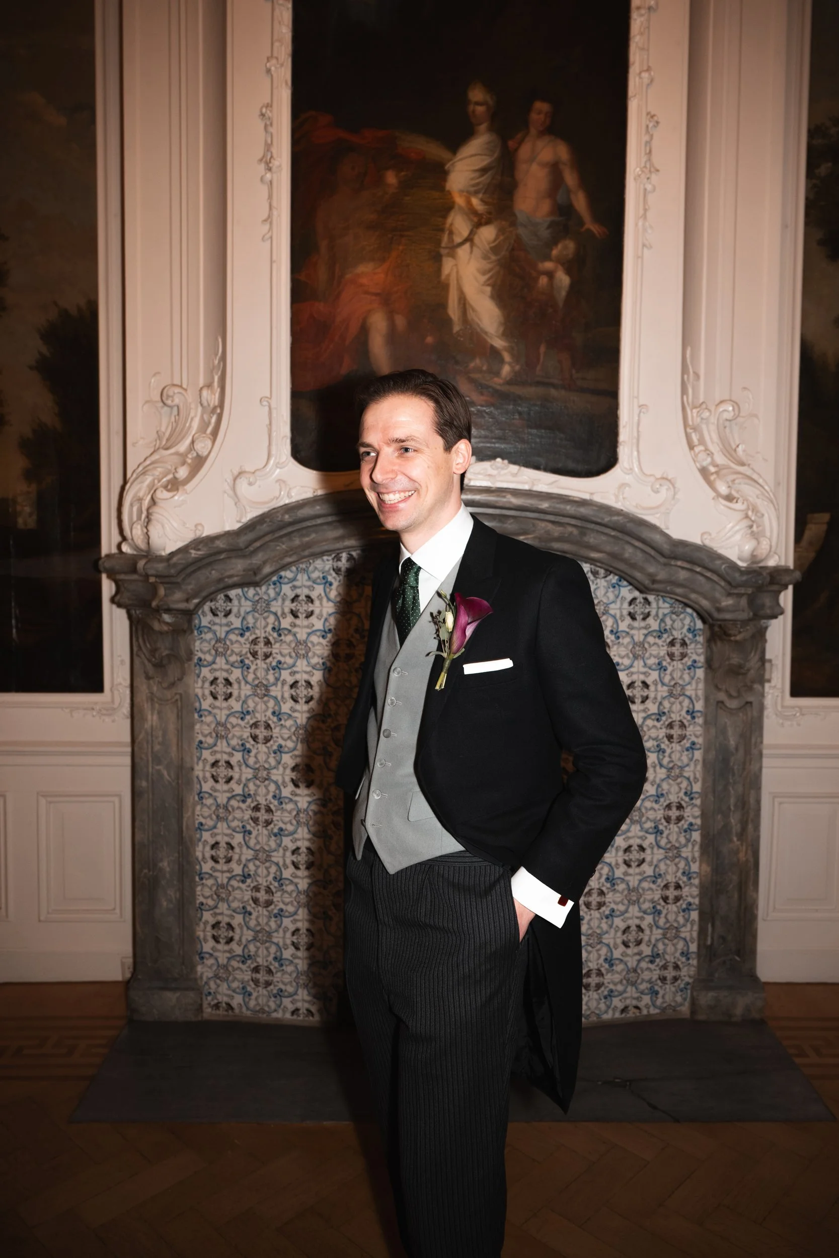 A smiling man in formal attire, including a black tuxedo with a gray vest and a white shirt, standing in front of a decorative fireplace with intricate detailing and a large painting or mural above it. The man has a dark green boutonniere pinned to h
