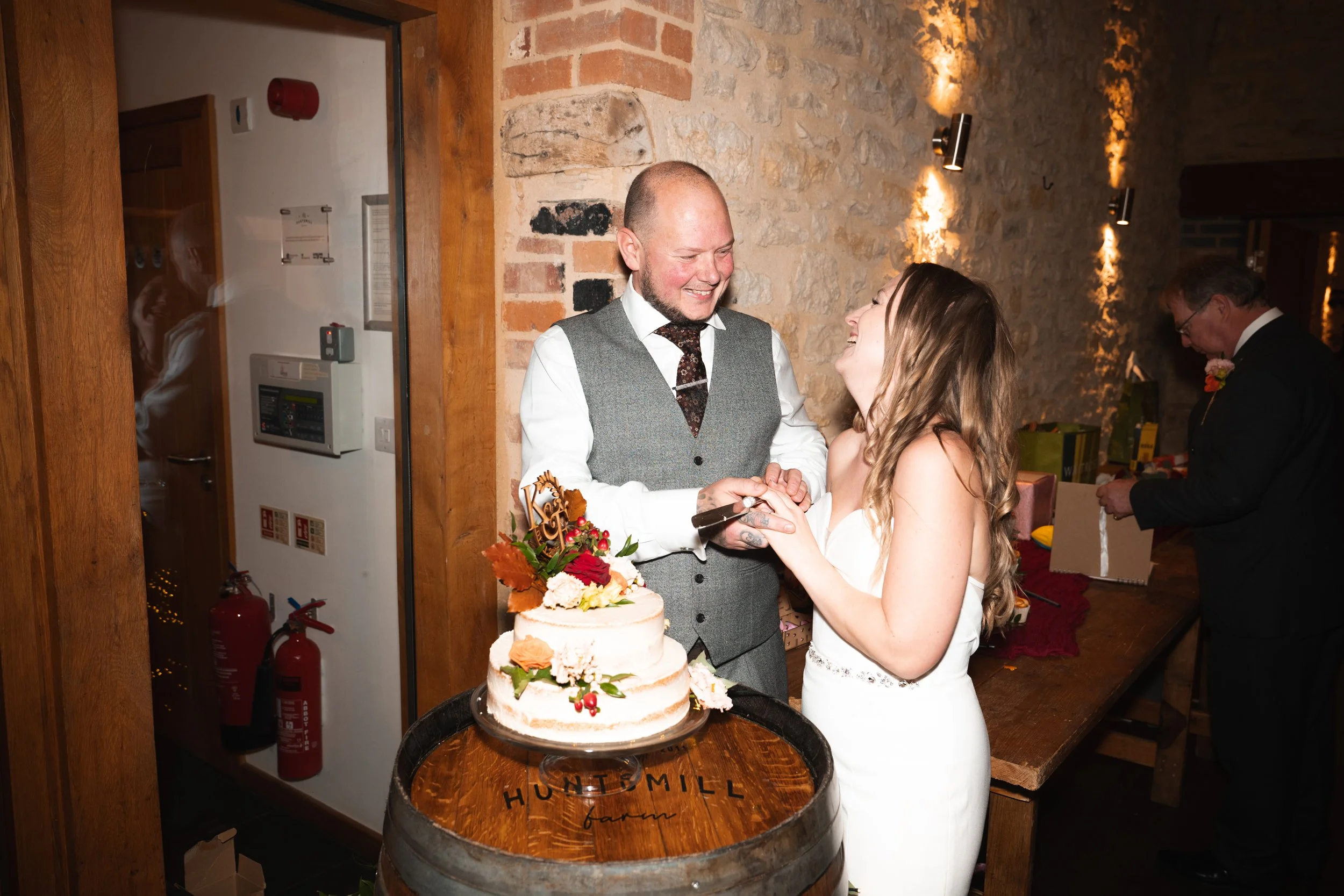 A wedding celebration with a bride and groom cutting a wedding cake together, smiling and holding hands, while a man looks on in the background. The scene takes place in a rustic venue with exposed brick and wooden accents.