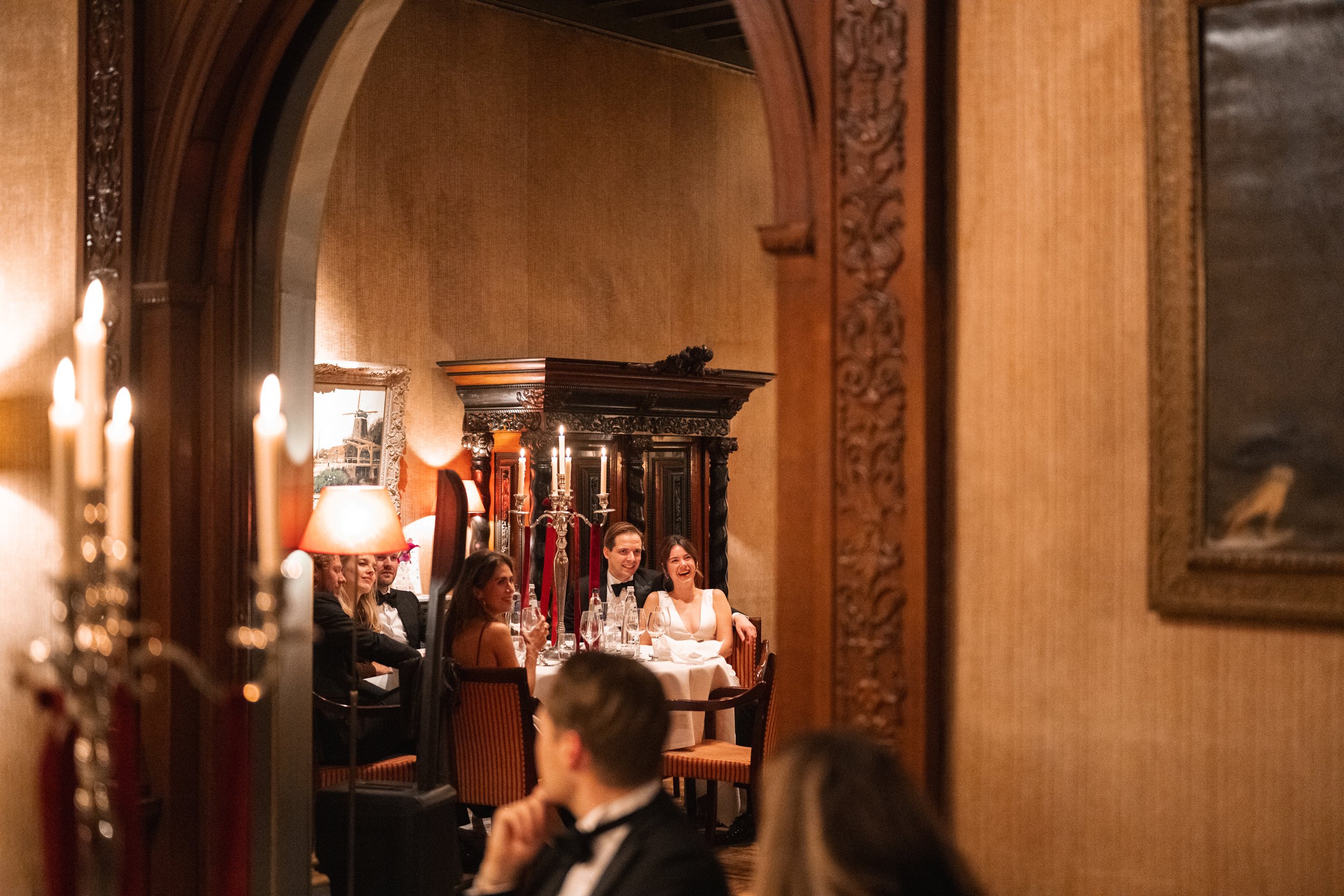 A group of people dressed formally at a dinner party, seen through an ornate doorway in a richly decorated room with warm lighting.