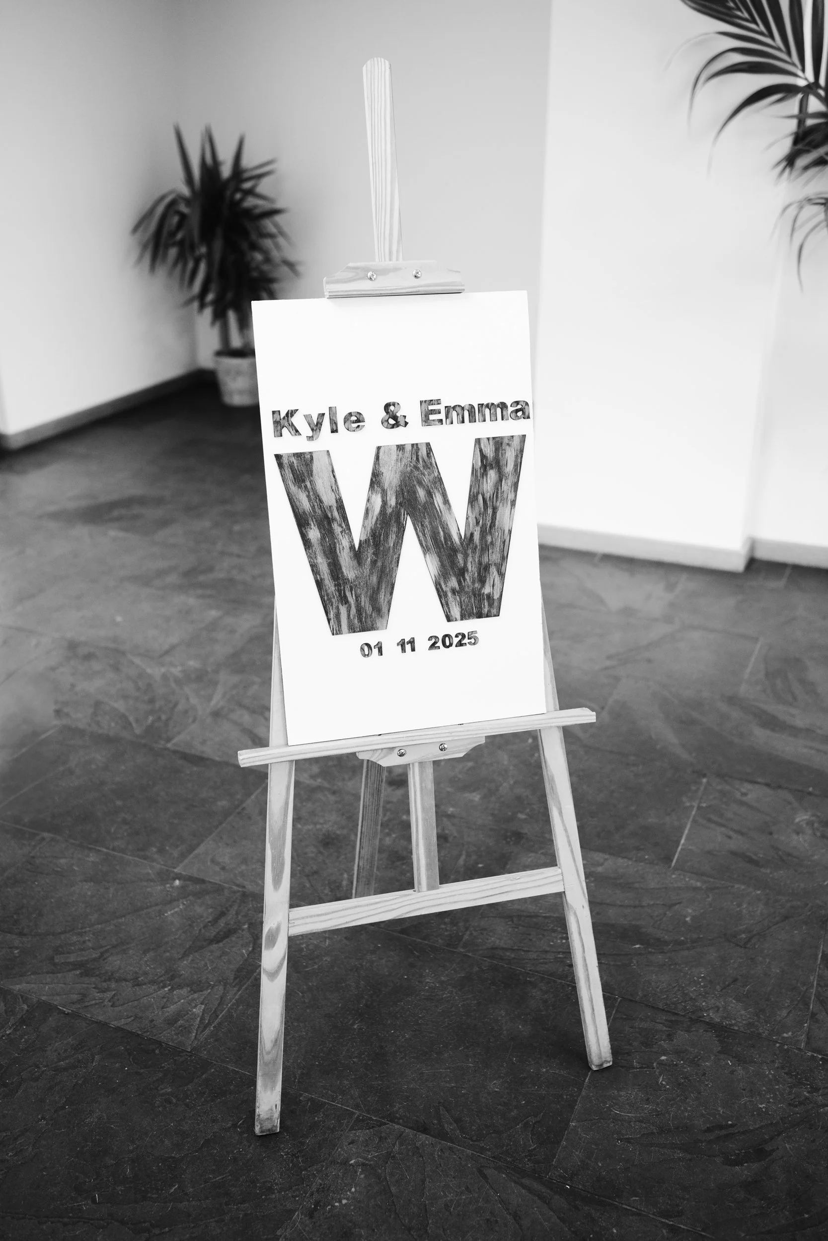 A wedding sign on an easel with the names Kyle and Emma, a large letter W, and the date 01 11 2025.
