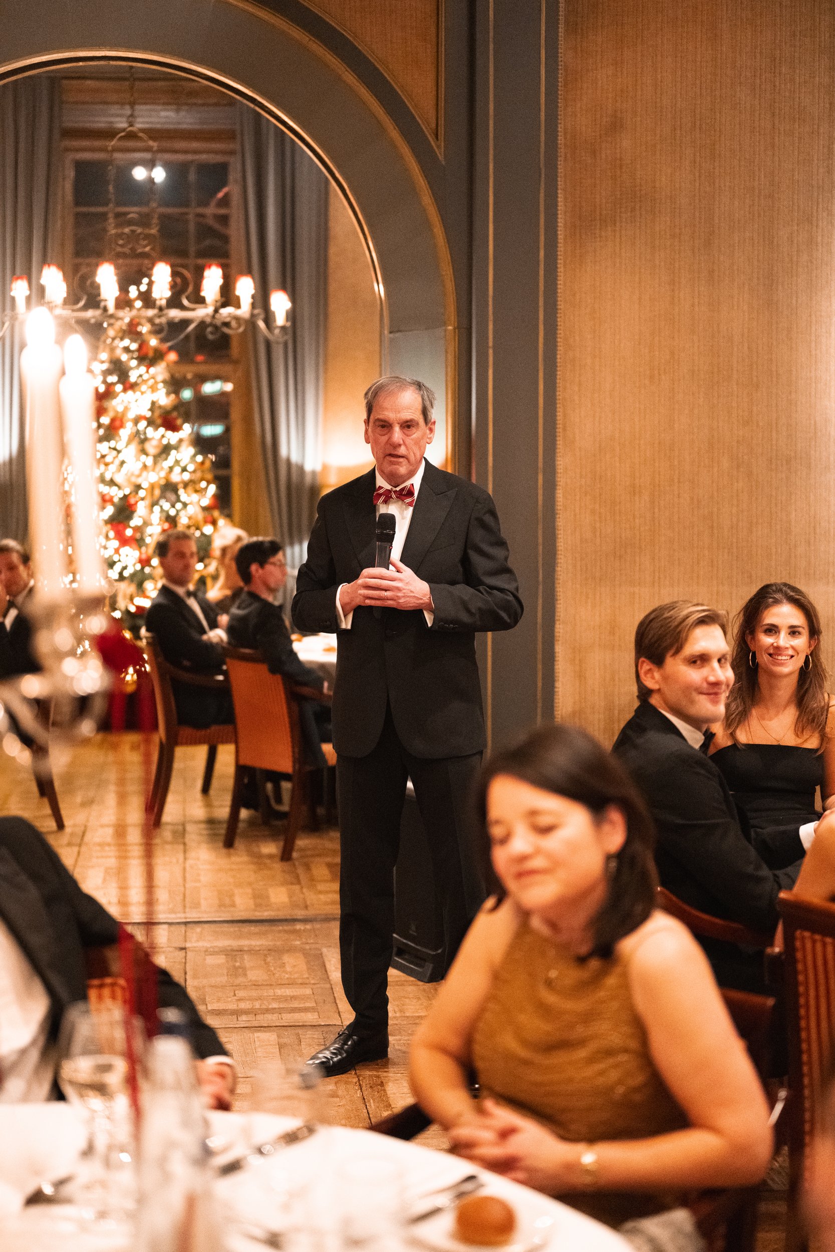 A man in a tuxedo holding a microphone speaking at a formal event, with guests seated at tables and a decorated Christmas tree in the background.