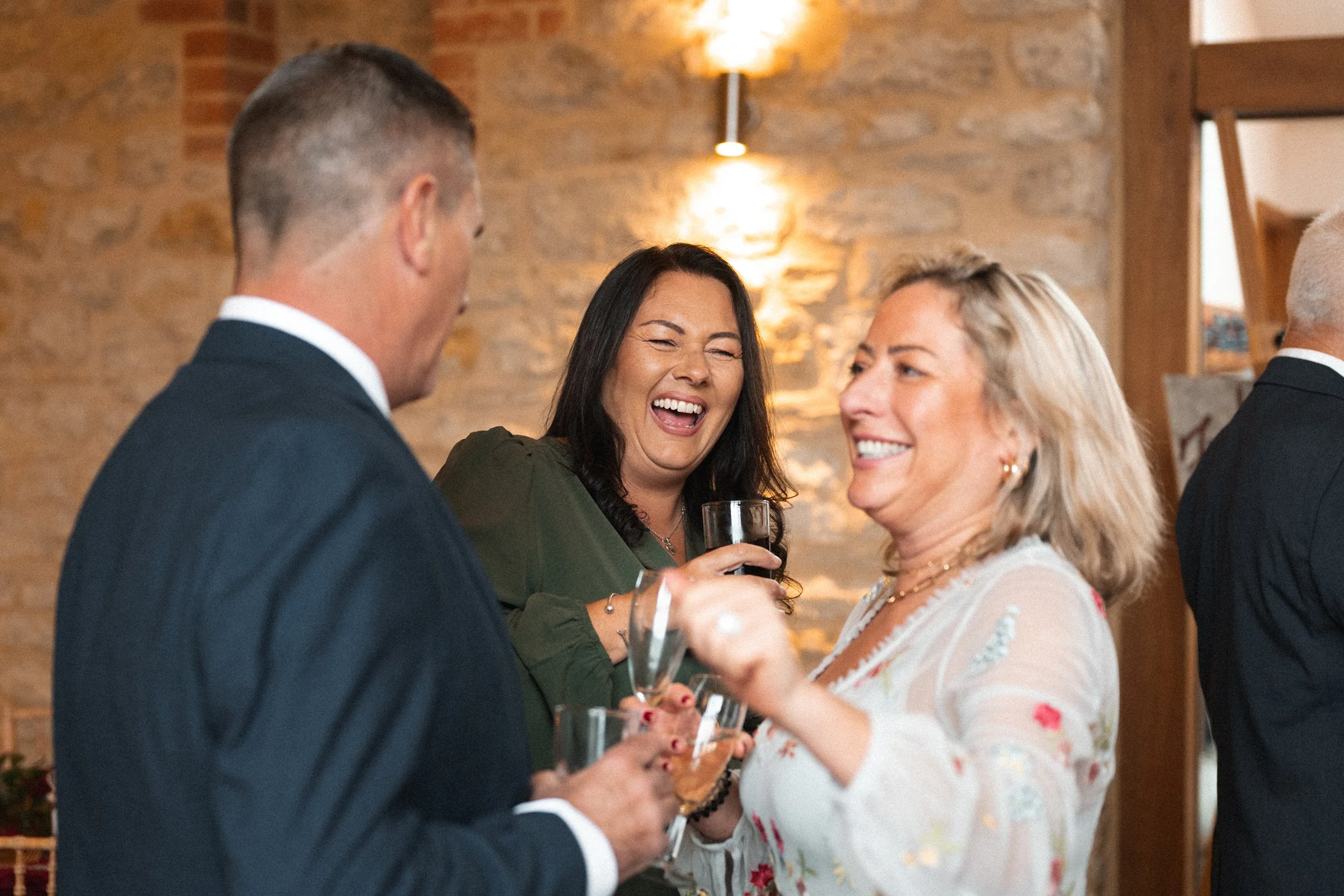 Four people at a social gathering, two women and two men, are talking and smiling, holding drinks in a warmly lit room with a brick wall background.