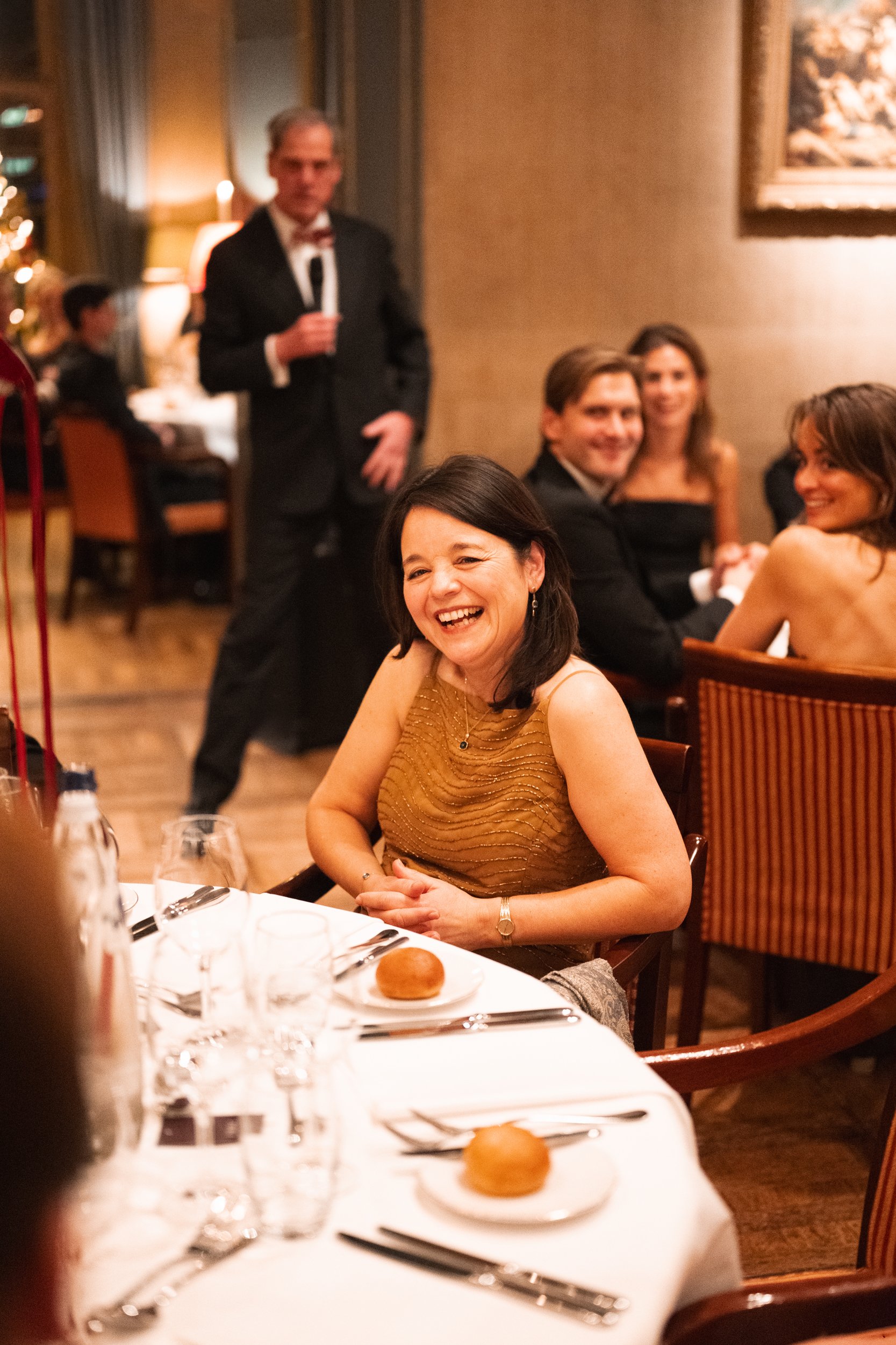 A woman with dark hair, wearing a gold sleeveless dress, sitting at a formal dinner table, smiling, with bread rolls, glasses, and silverware on the table. In the background, other guests are seated and a waiter in a black suit with a bow tie stands 