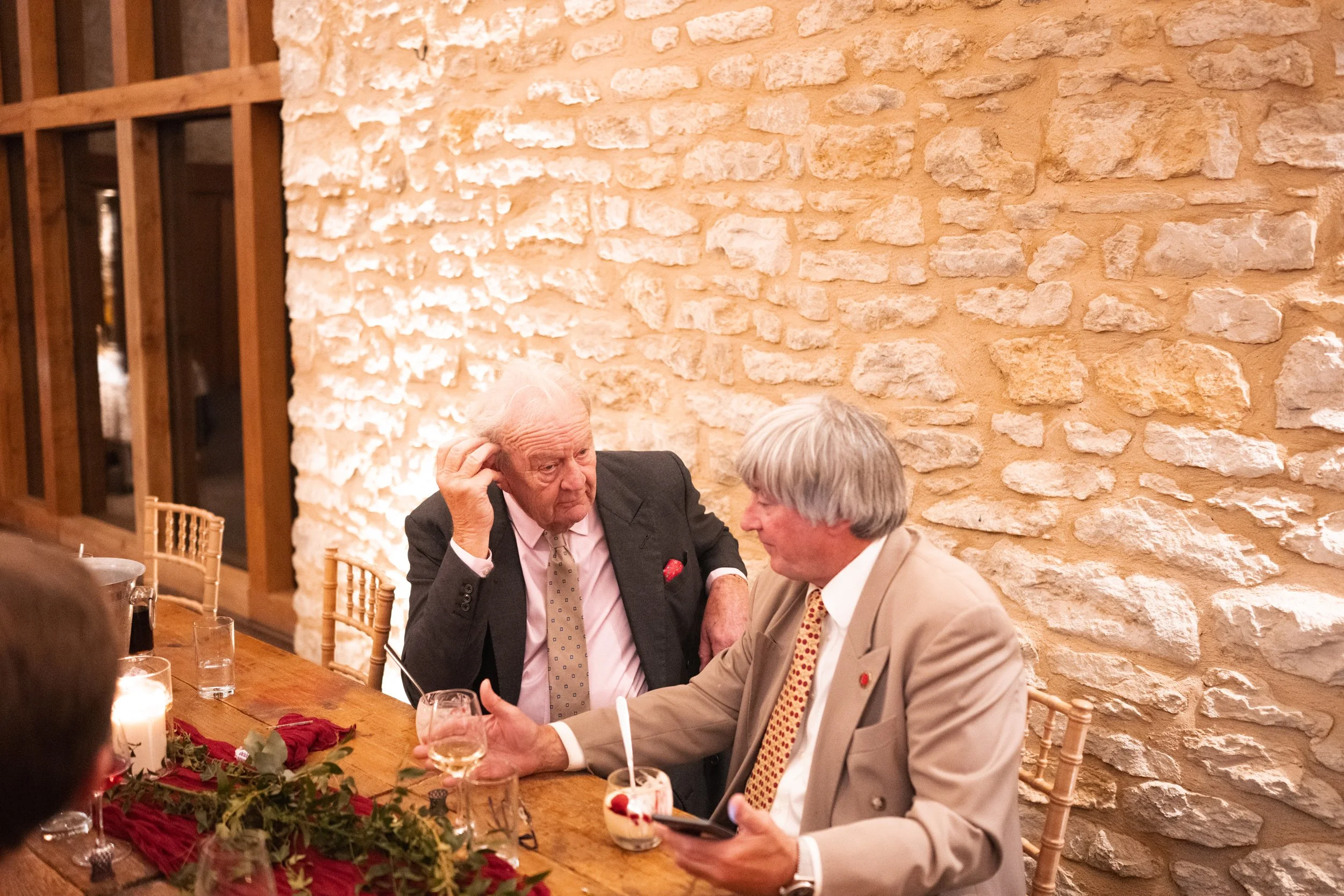 Two elderly men in suits having a conversation at a decorated wooden table in a rustic-style room with white stone walls. One man is touching his head, and the other is holding a drink and a smartphone.