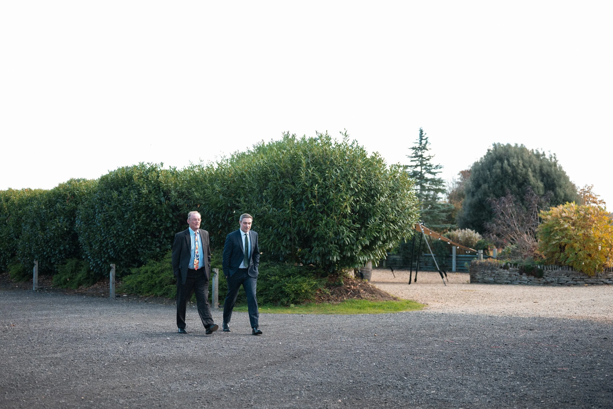 Two men in suits walking outdoors near large green bushes and trees, with a swing set and playground equipment in the background.