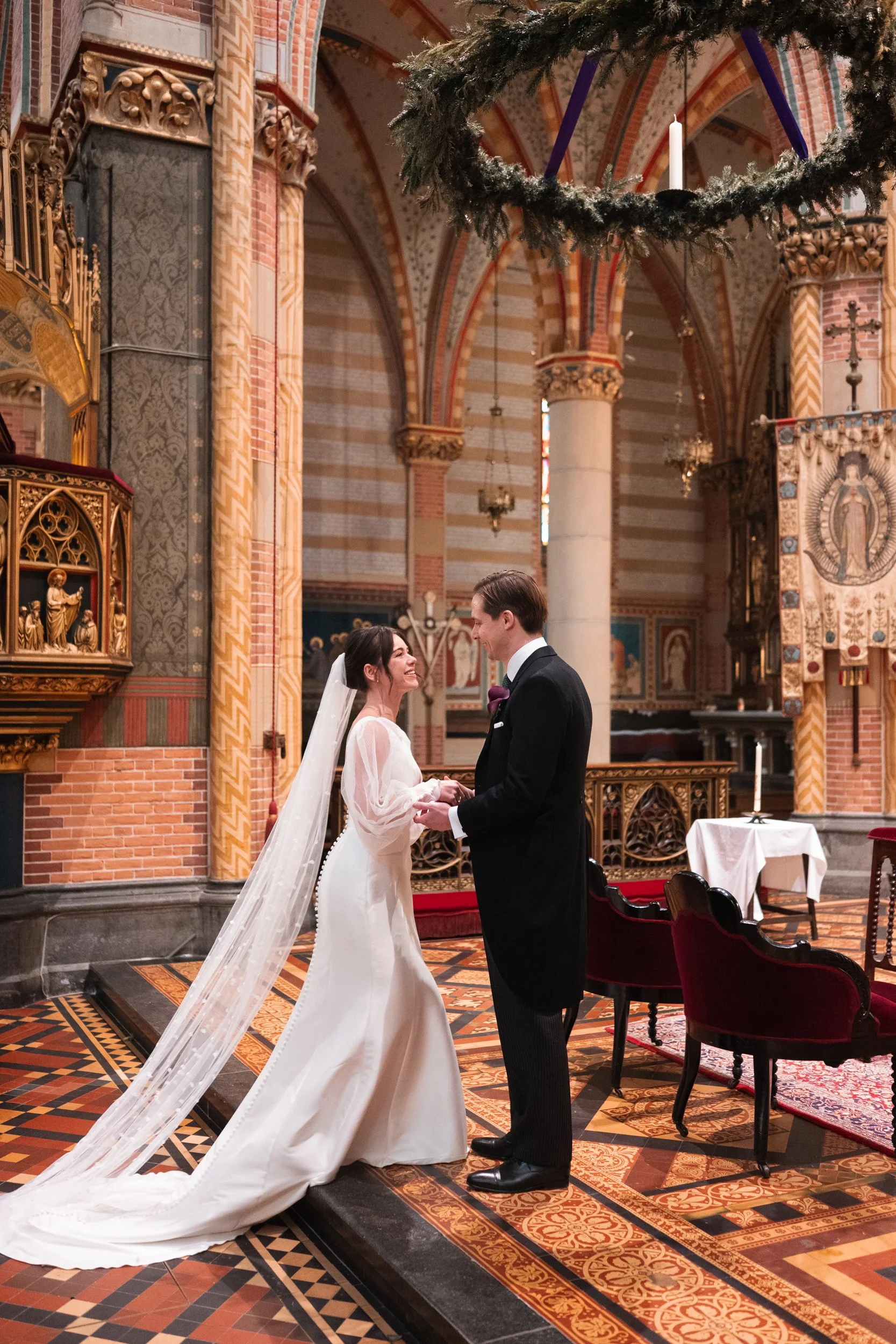 A bride and groom are holding hands during their wedding ceremony inside a church with ornate decorations, stained glass windows, and a large Christmas wreath hanging above.