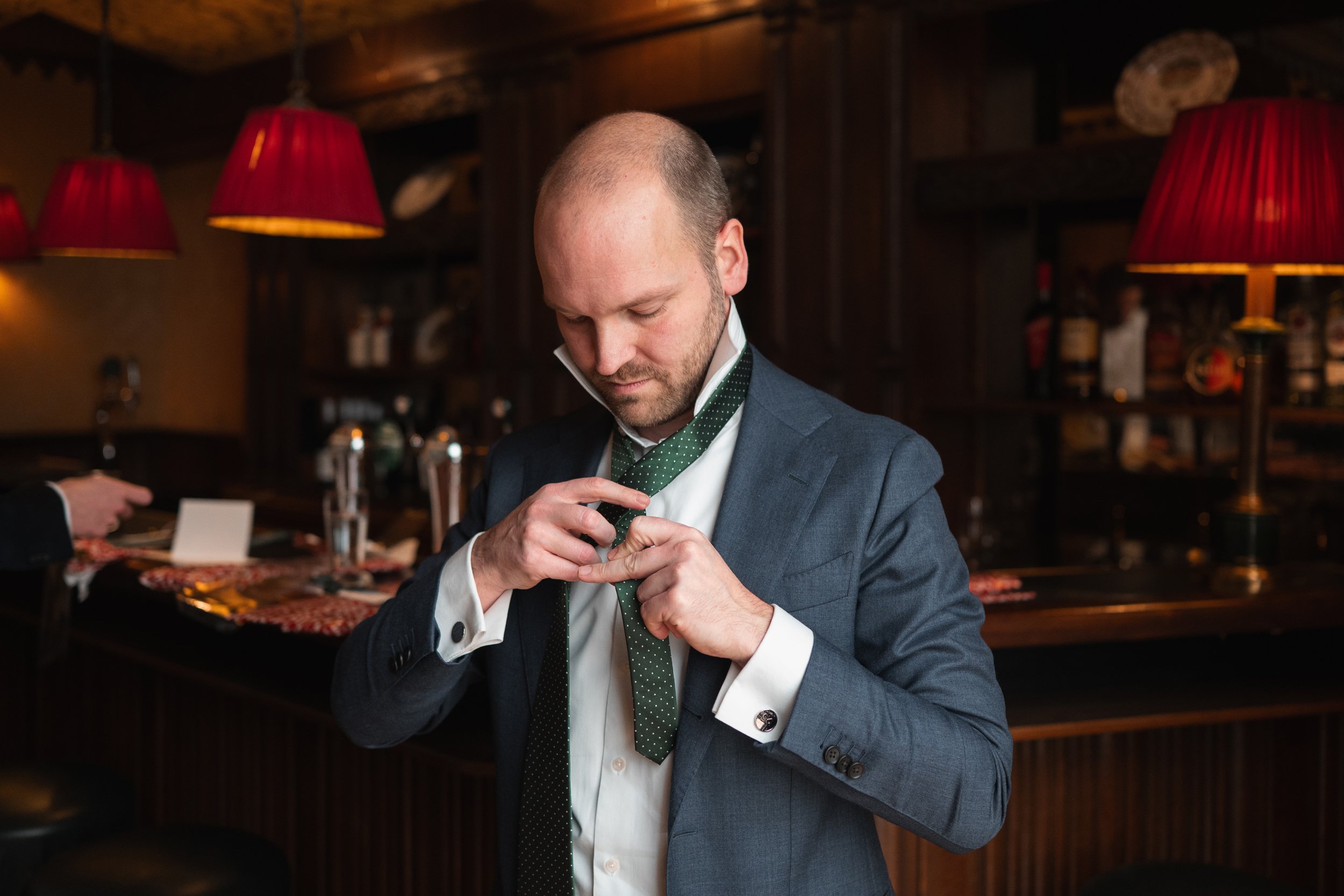 A man in a blue suit and white shirt is adjusting his green polka dot tie inside a dimly lit bar or restaurant with red lampshades.