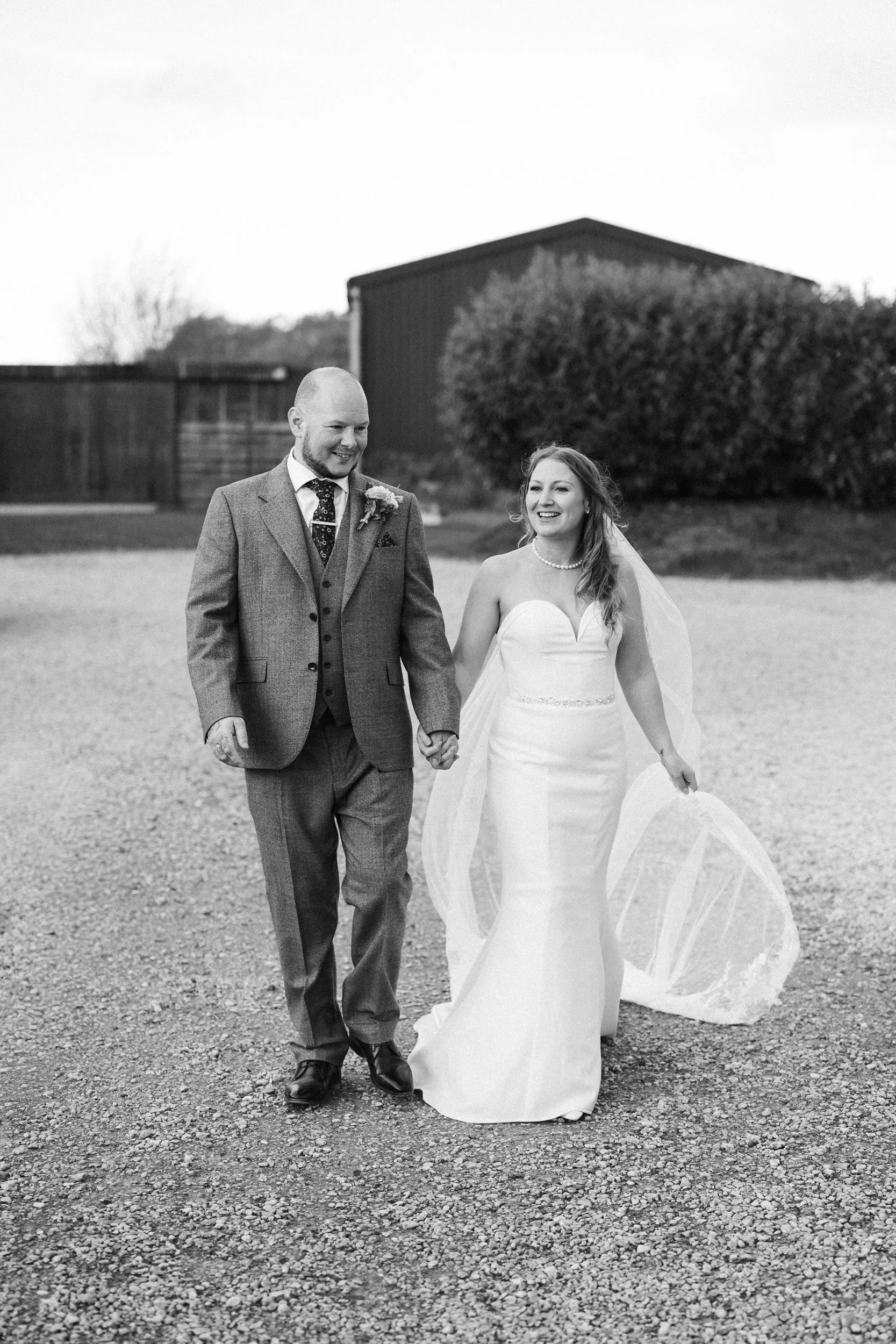 A black and white photo of a newlywed couple holding hands and walking outdoors, smiling, with the bride in a strapless white wedding gown and the groom in a gray suit with a tie.