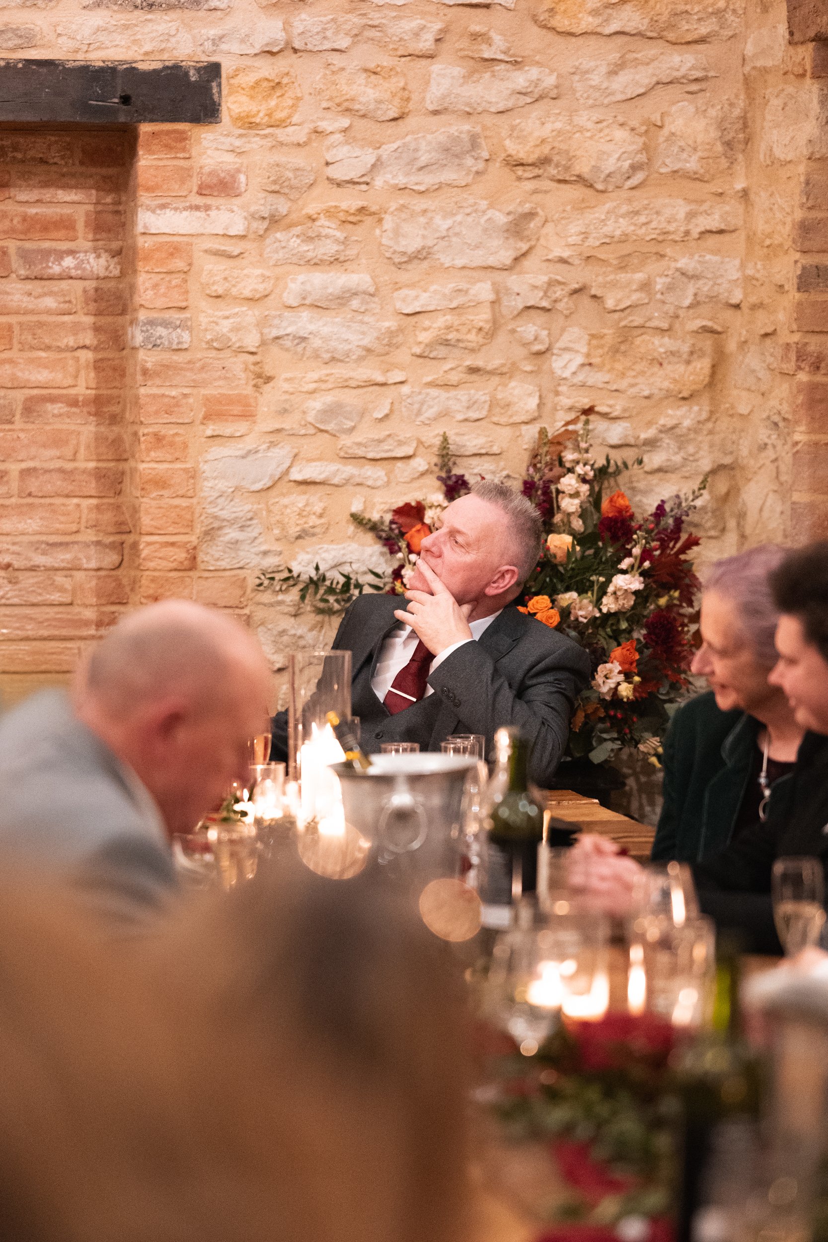 A man in a suit sitting at a dining table with a flower arrangement behind him, appearing thoughtful or contemplative, during a formal dinner or event.