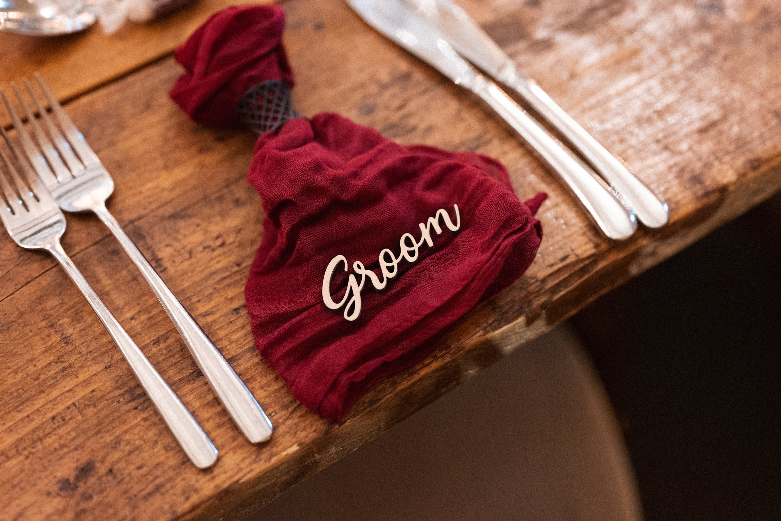 A wooden dining table set with three forks on the left, a red cloth napkin with a 'Groom' sign in the center, and a knife and spoon on the right.