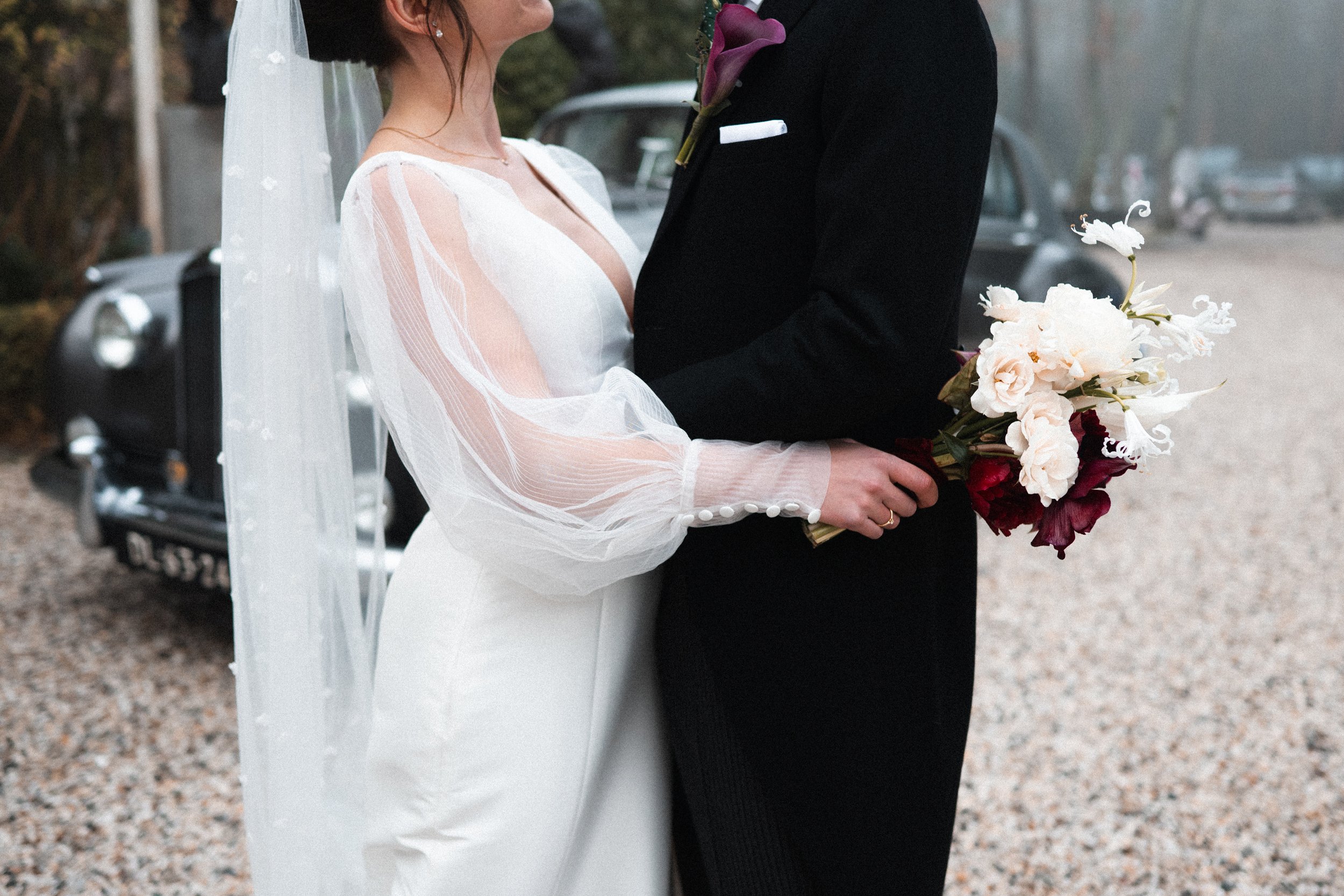 Bride and groom holding hands at their wedding, with the bride wearing a white dress with sheer, puffed sleeves and a veil, and the groom in a black suit holding a bouquet of white and dark red flowers, with vintage cars in the background.