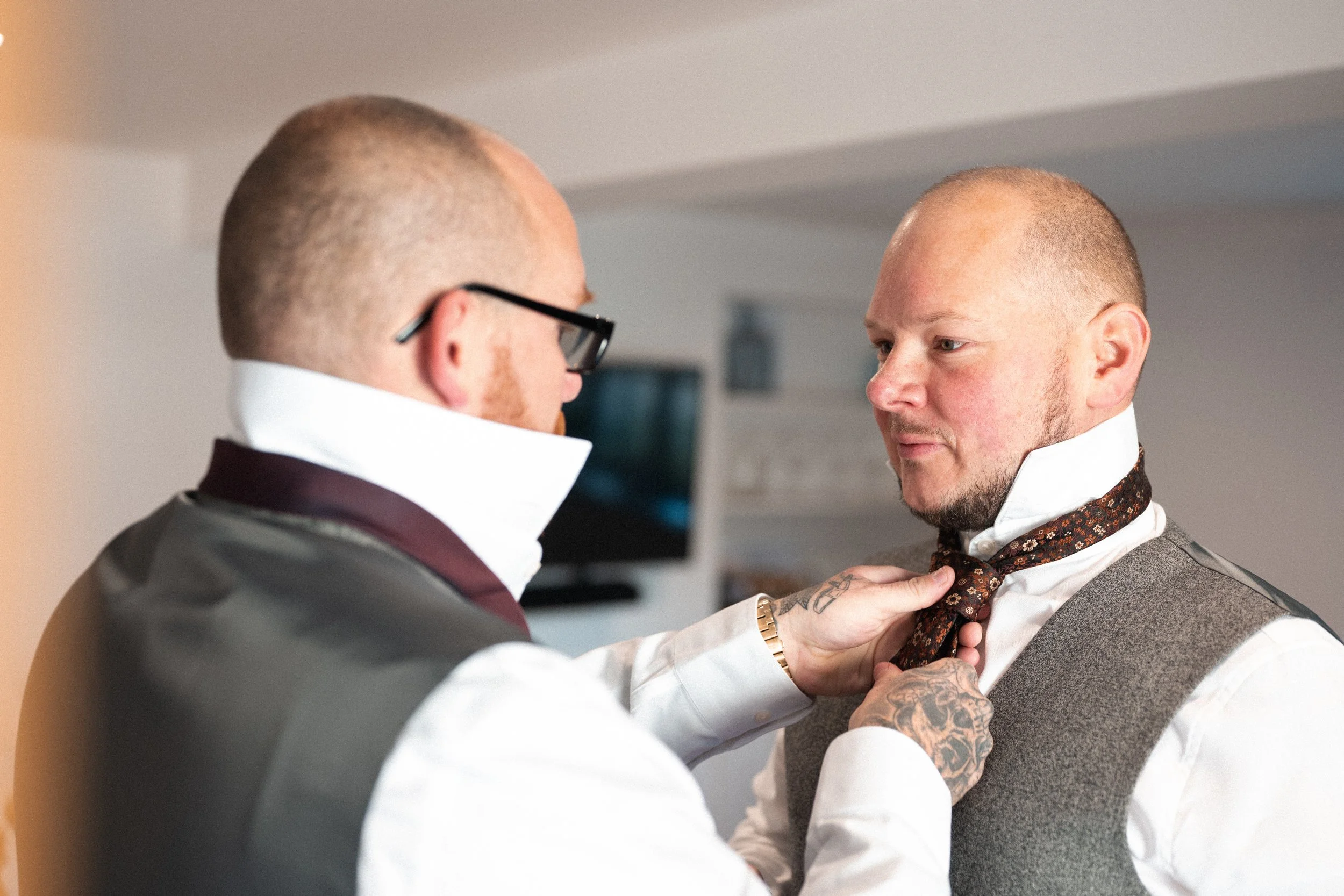 Two men with shaved heads, one with glasses, are adjusting a tie on the other man in a well-lit room. The man on the right is wearing a white shirt and a gray vest, while the man on the left is wearing a white shirt with a high collar and a dark vest