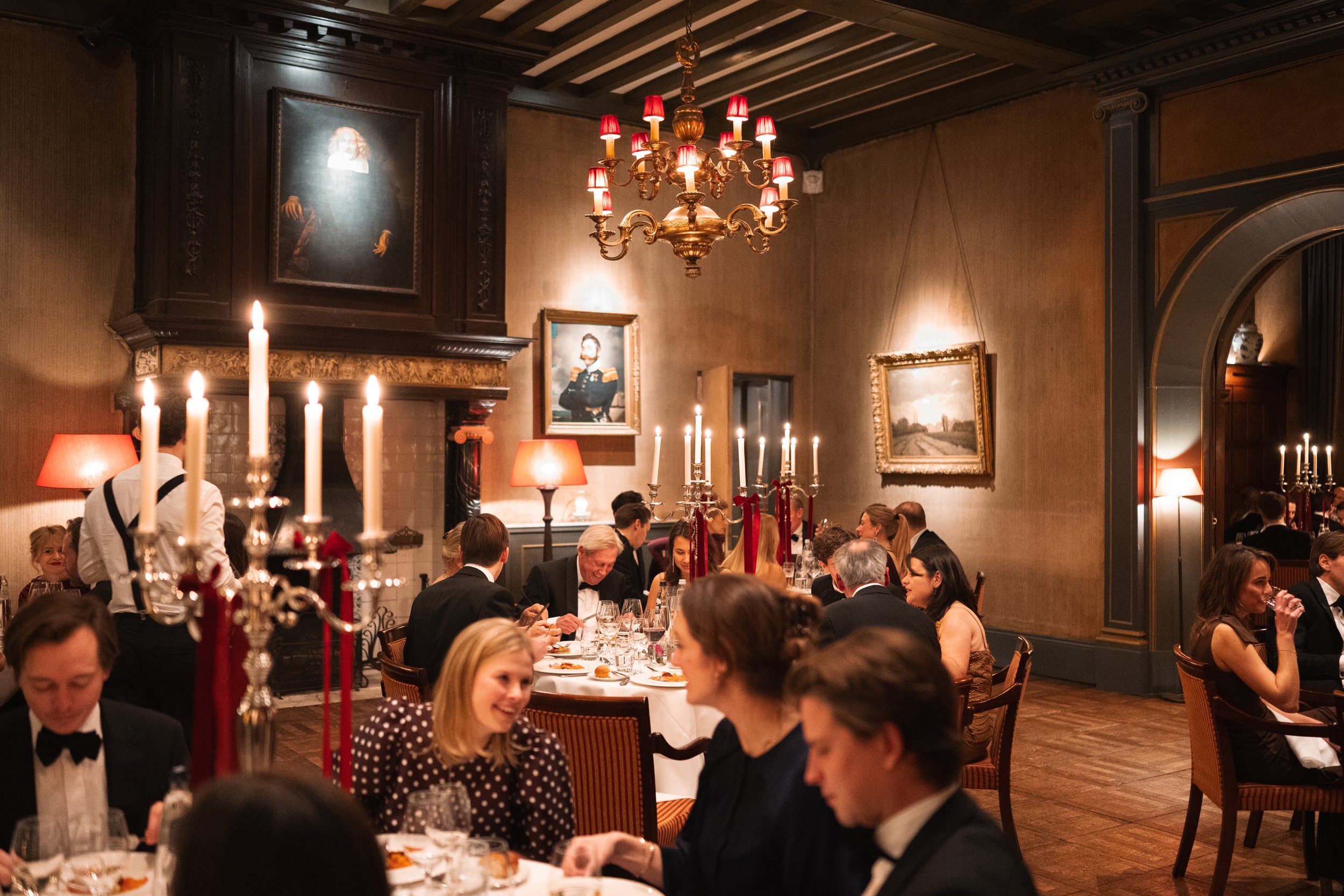 Guests dining at a formal dinner event in an elegant room with warm lighting, paintings on the walls, and a large chandelier, with some tables featuring candelabras and red ribbons.