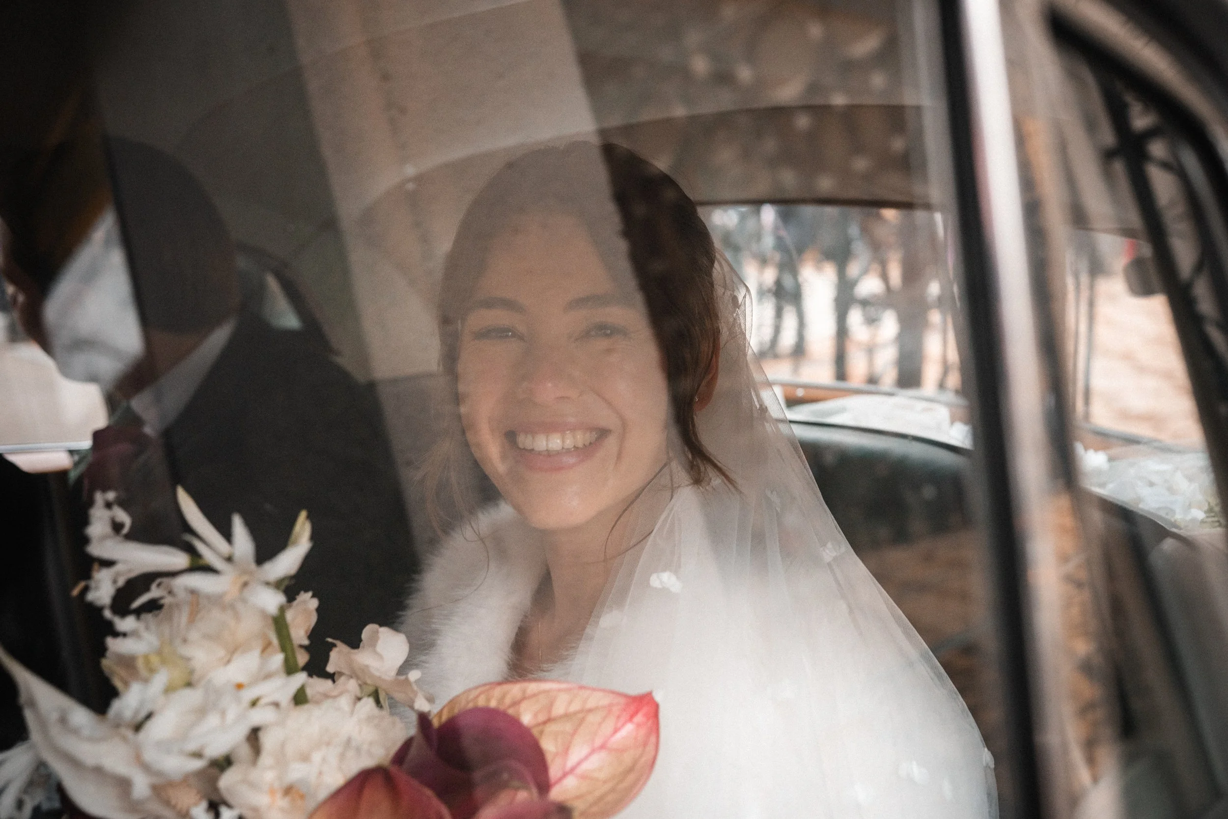 A smiling woman in bridal attire sitting inside a vehicle with a veil over her face, holding a bouquet of flowers, with trees visible outside through the window.