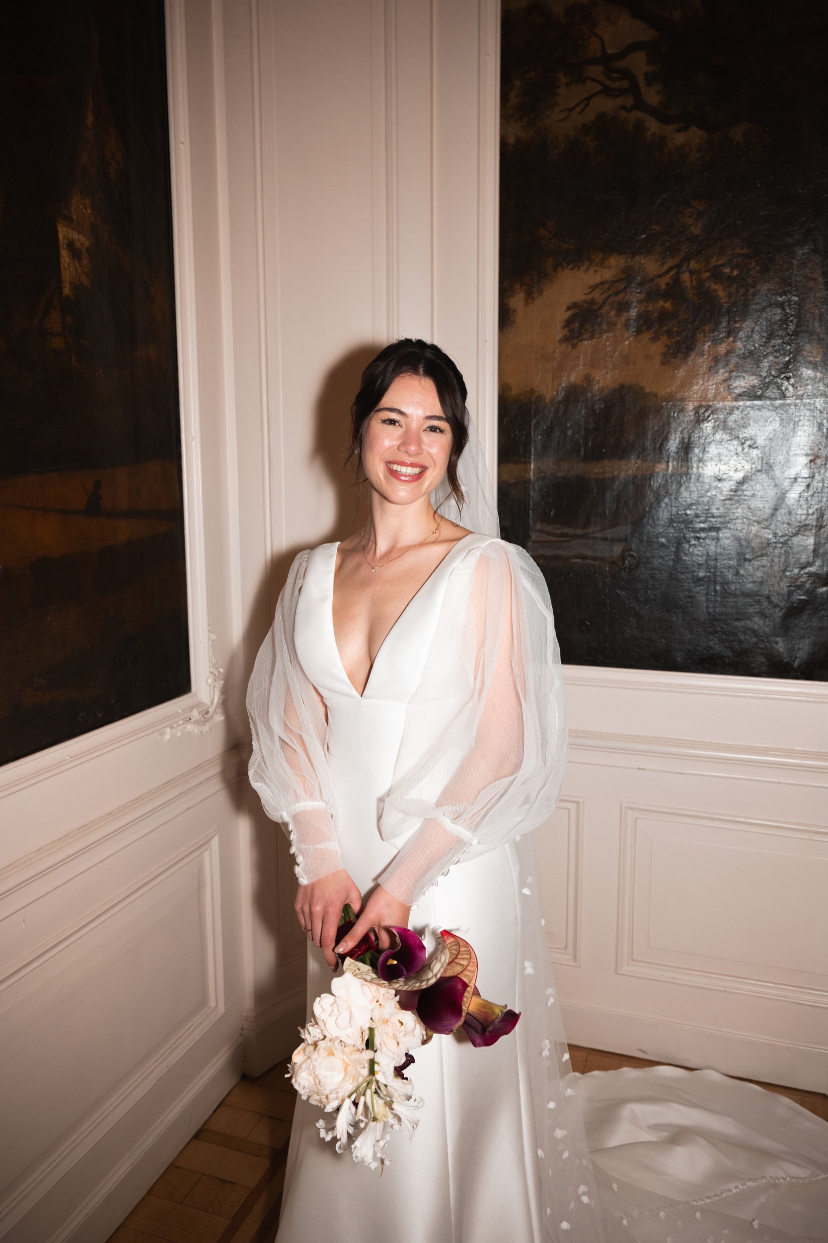 A woman in a white wedding dress holding a bouquet, standing indoors near a wall with dark paintings.