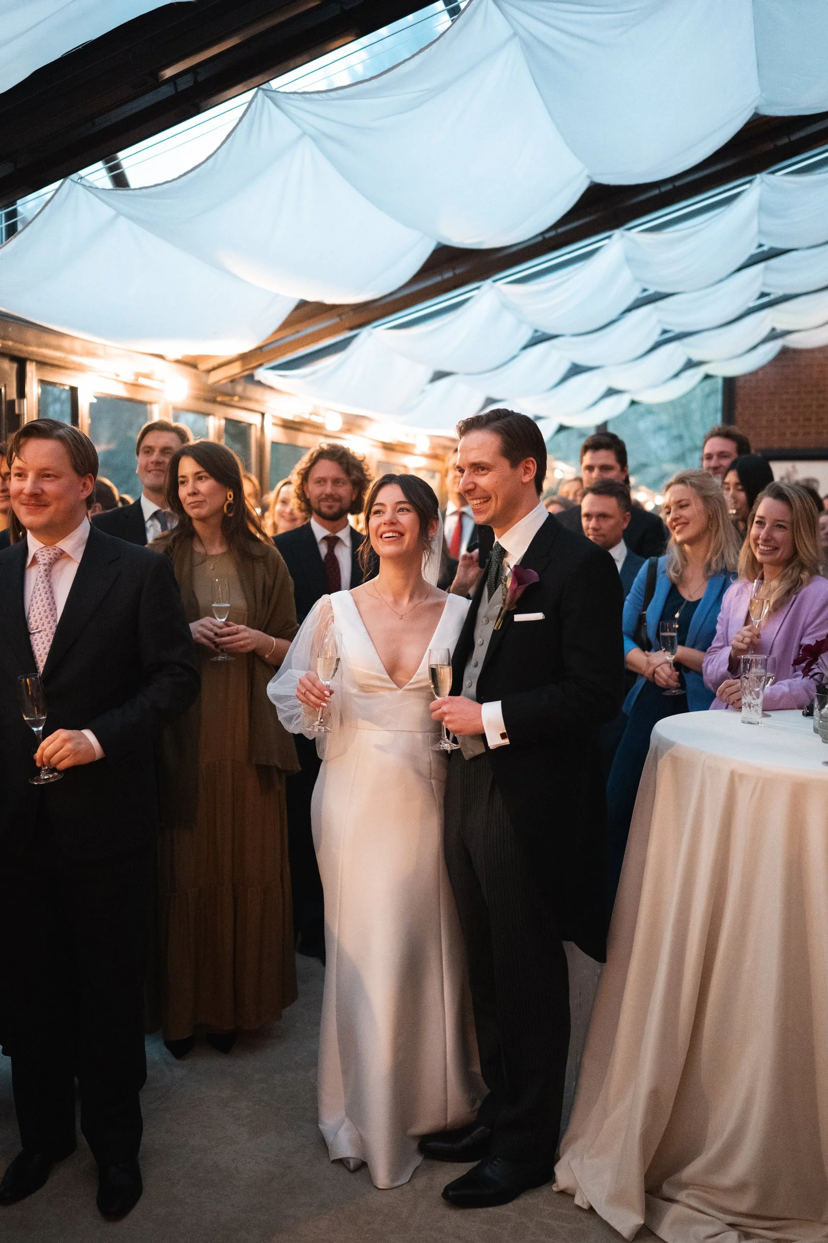 Bride and groom at wedding reception holding champagne glasses, surrounded by happy guests, decorated venue with white fabric ceiling drapes.