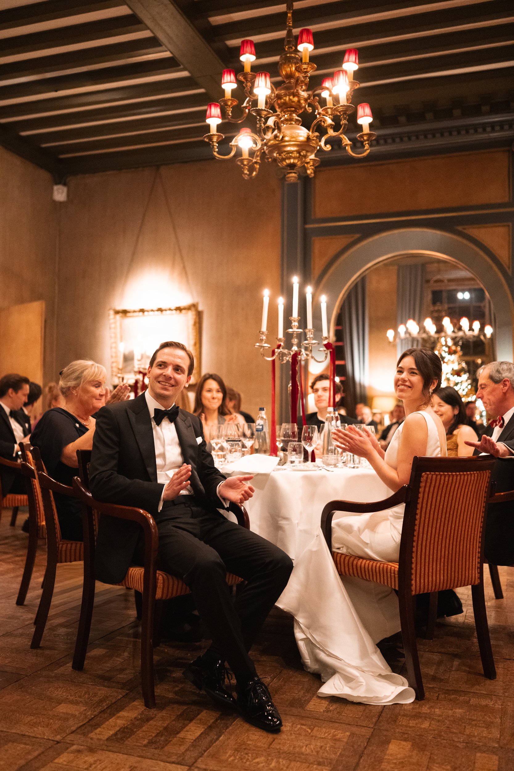 People sitting at a decorated dinner table during a formal event or celebration, with a chandelier overhead and warm lighting.
