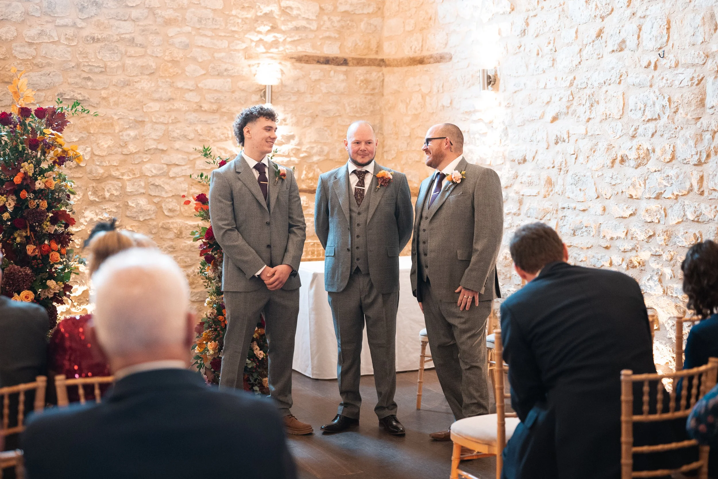 Three men in gray suits standing in front of an audience at a wedding ceremony inside a rustic stone-walled room. One man is smiling while the other two look at him, with floral decorations visible on the sides.
