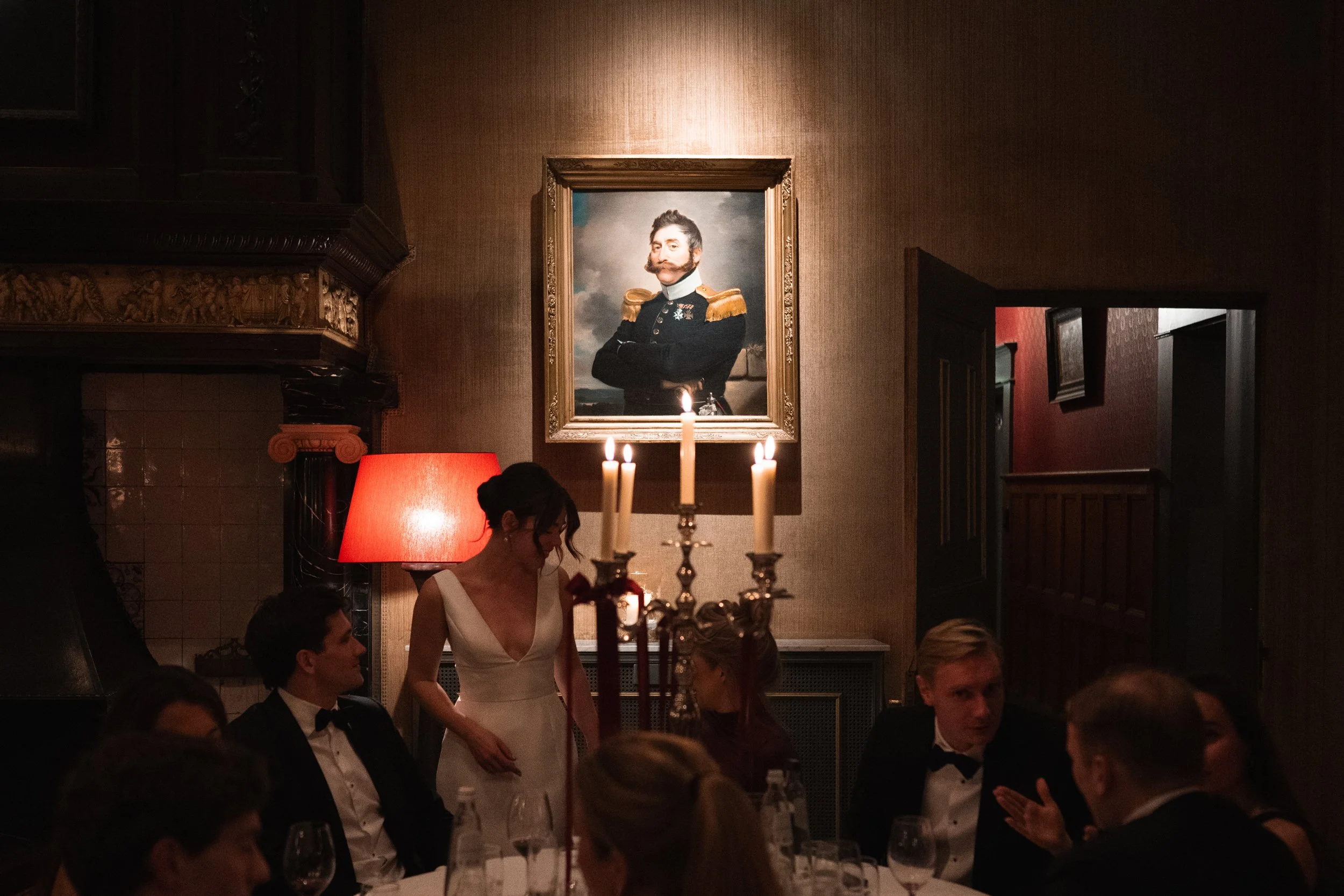A group of people dressed in formal attire dining at a restaurant with a woman in a white dress standing and talking among them. There is a candlestick holder with lit candles on the table, and a framed portrait of a man in a naval uniform hangs on t