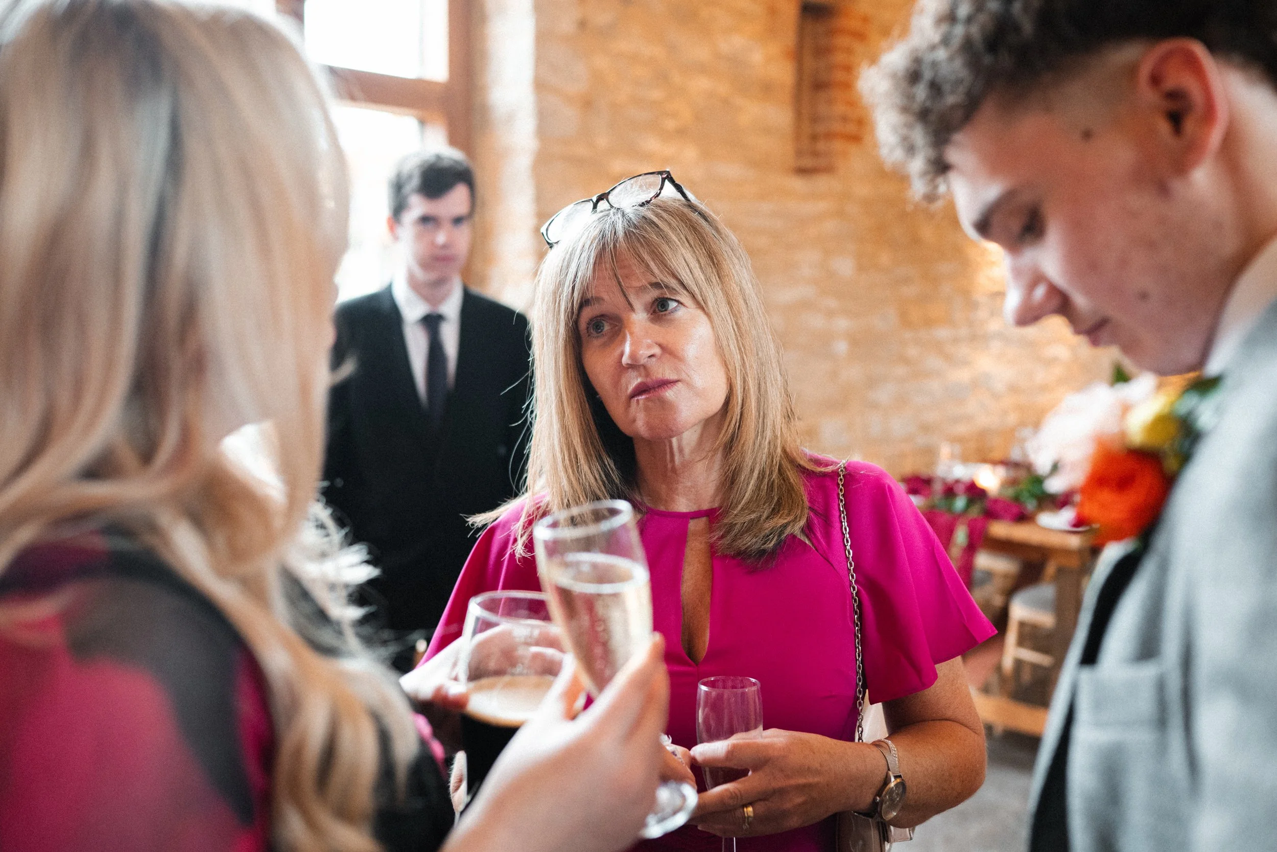 Group of people socializing at a party with drinks, brick wall in the background, woman in a pink dress talking and holding a glass of champagne.