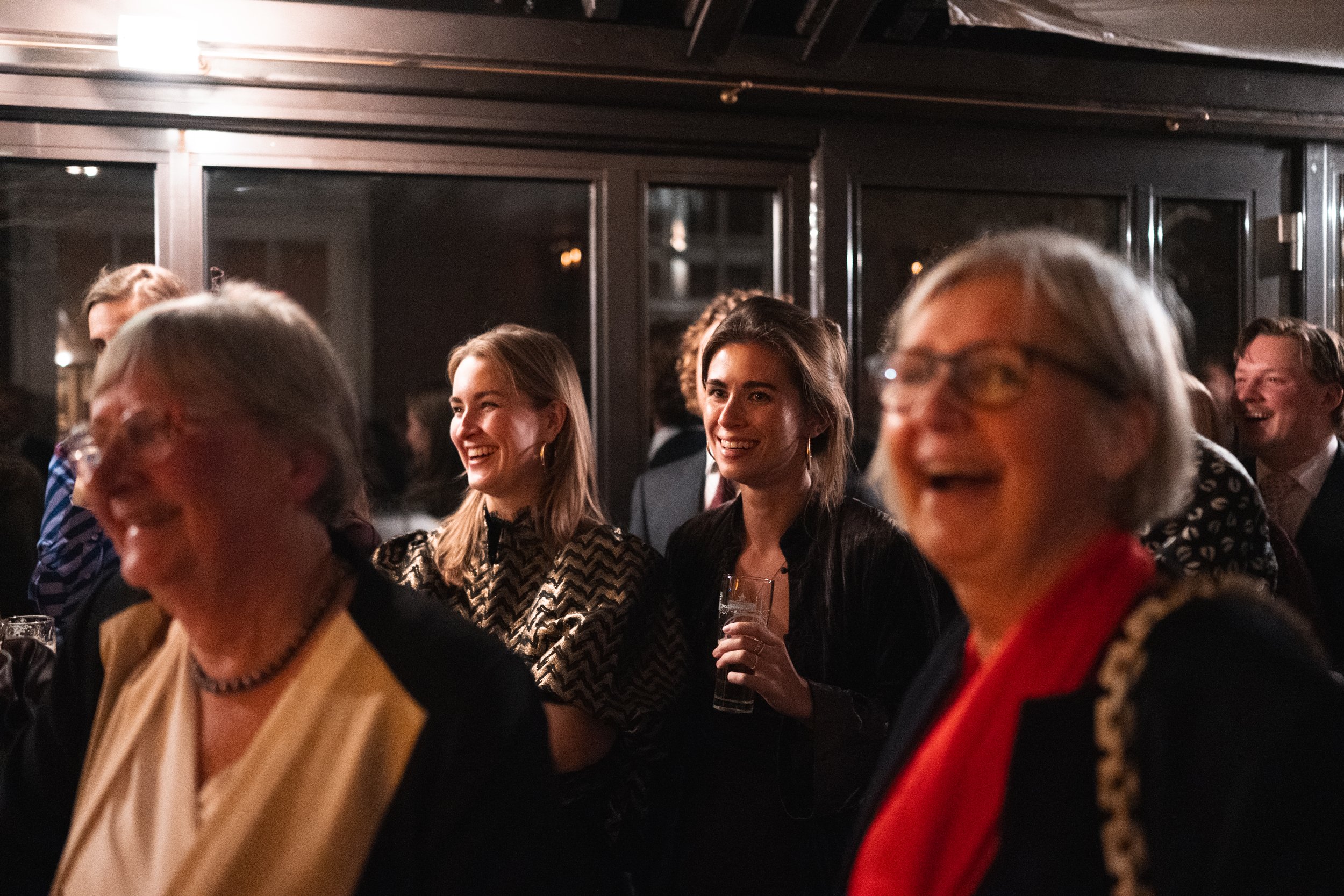 People enjoying a social gathering at night. Women are smiling, with one holding a glass of drink. The atmosphere is lively and cheerful.
