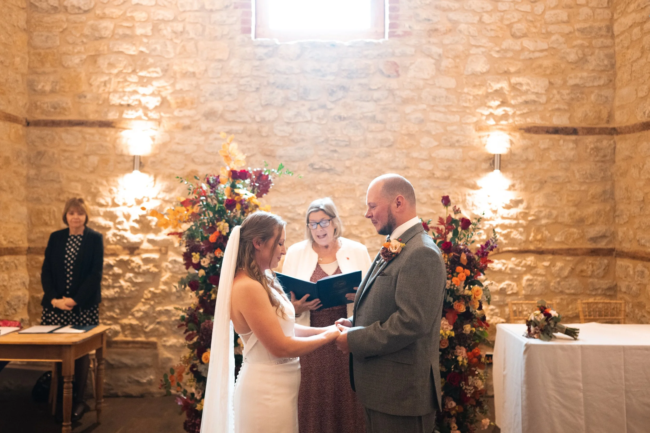 A bride and groom holding hands and exchanging vows during a wedding ceremony in a rustic stone-walled venue, with a female officiant and colorful floral arrangements behind them.