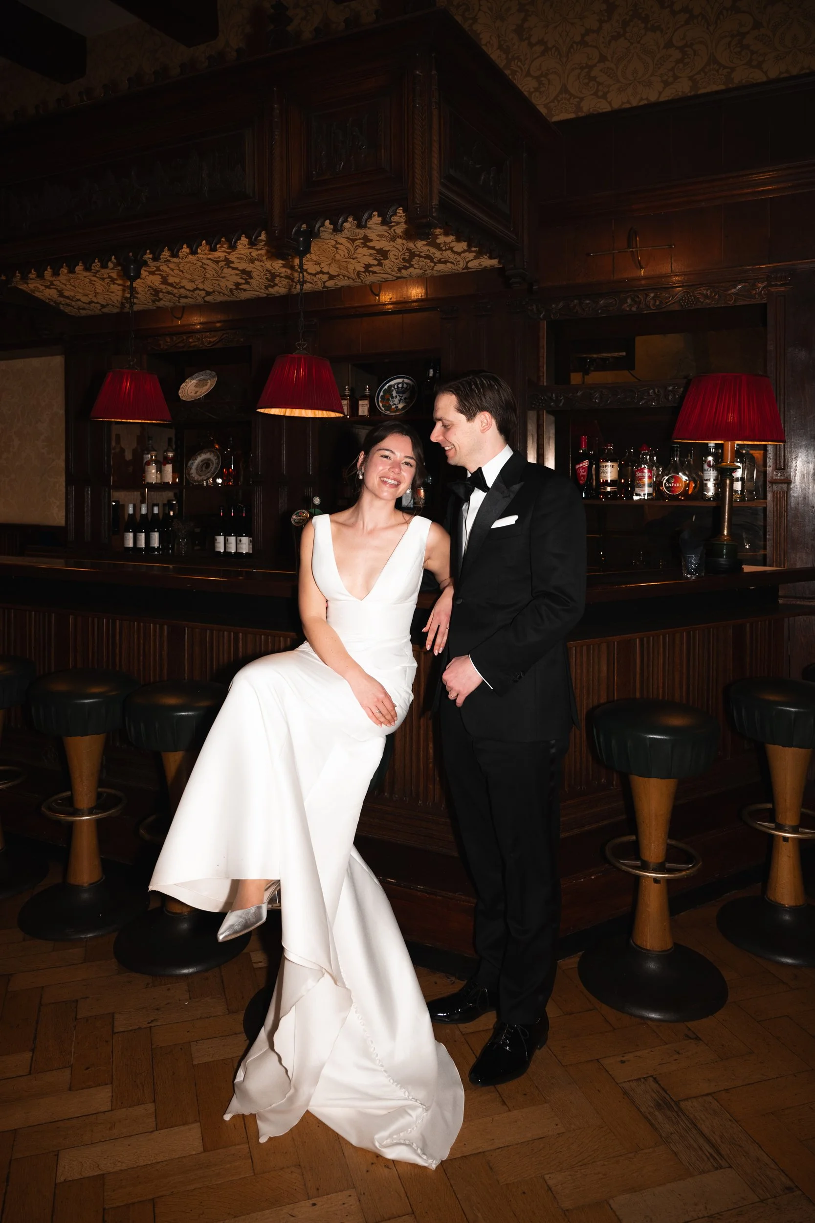 A bride in a white wedding gown sitting on a bar stool, smiling, with a groom in a black tuxedo standing beside her, in a dimly lit bar with wooden decor and red lampshades.