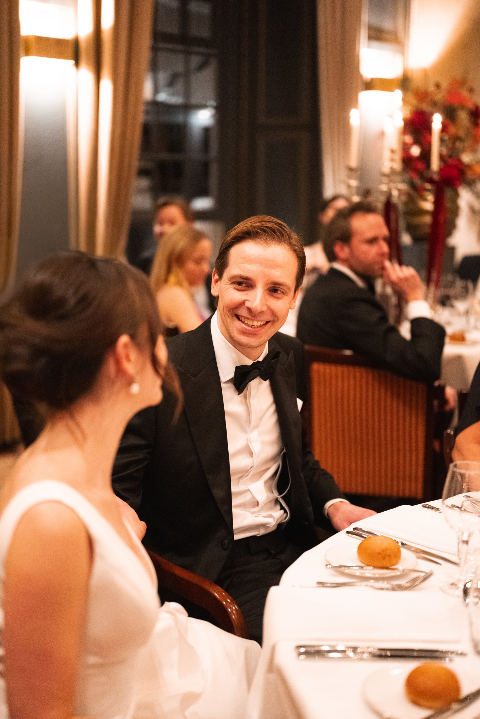 A man dressed in a tuxedo smiling and talking to a woman in a white dress at a formal dinner event.