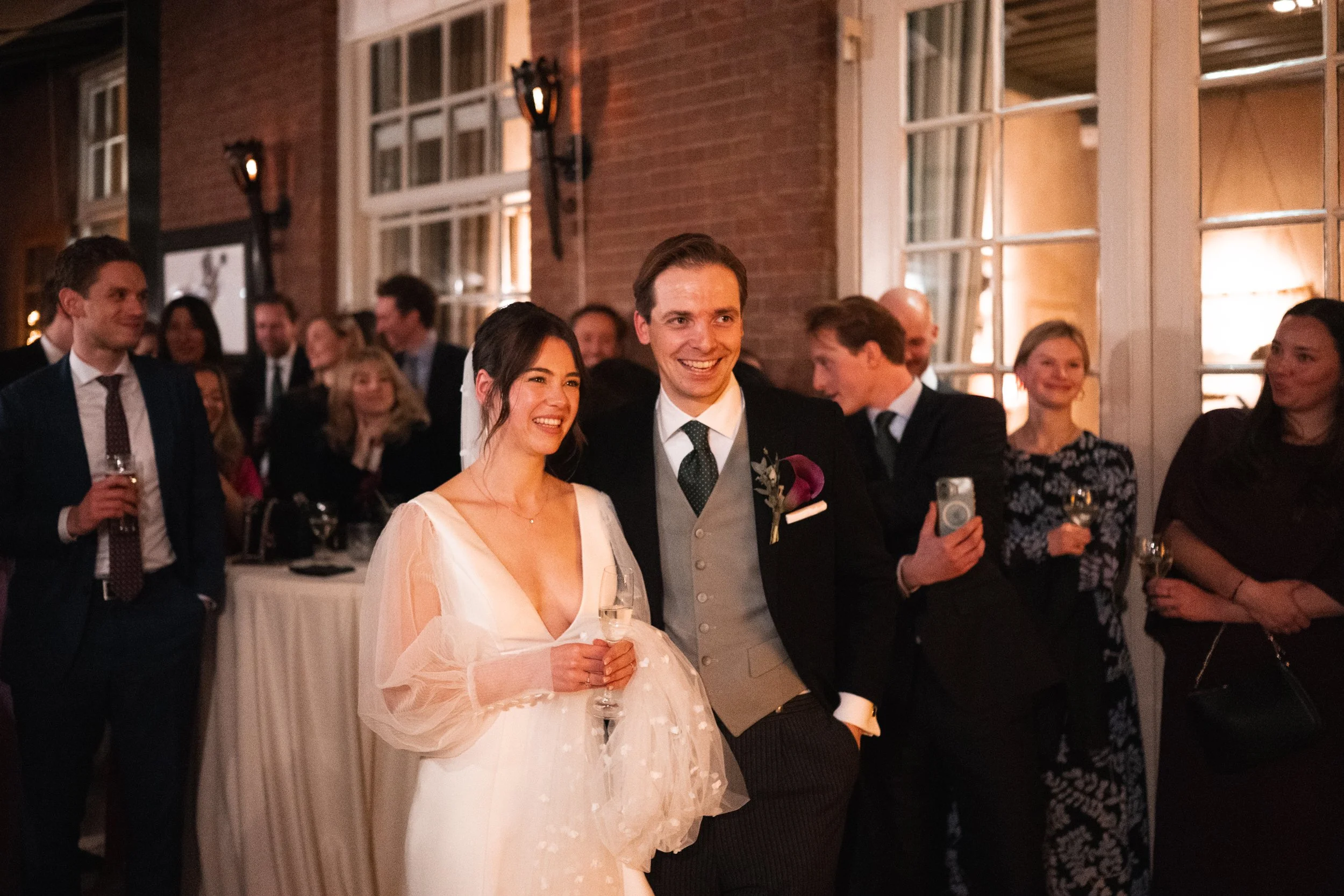 Bride and groom smiling at a wedding reception surrounded by guests, with some holding drinks and one taking a photo.
