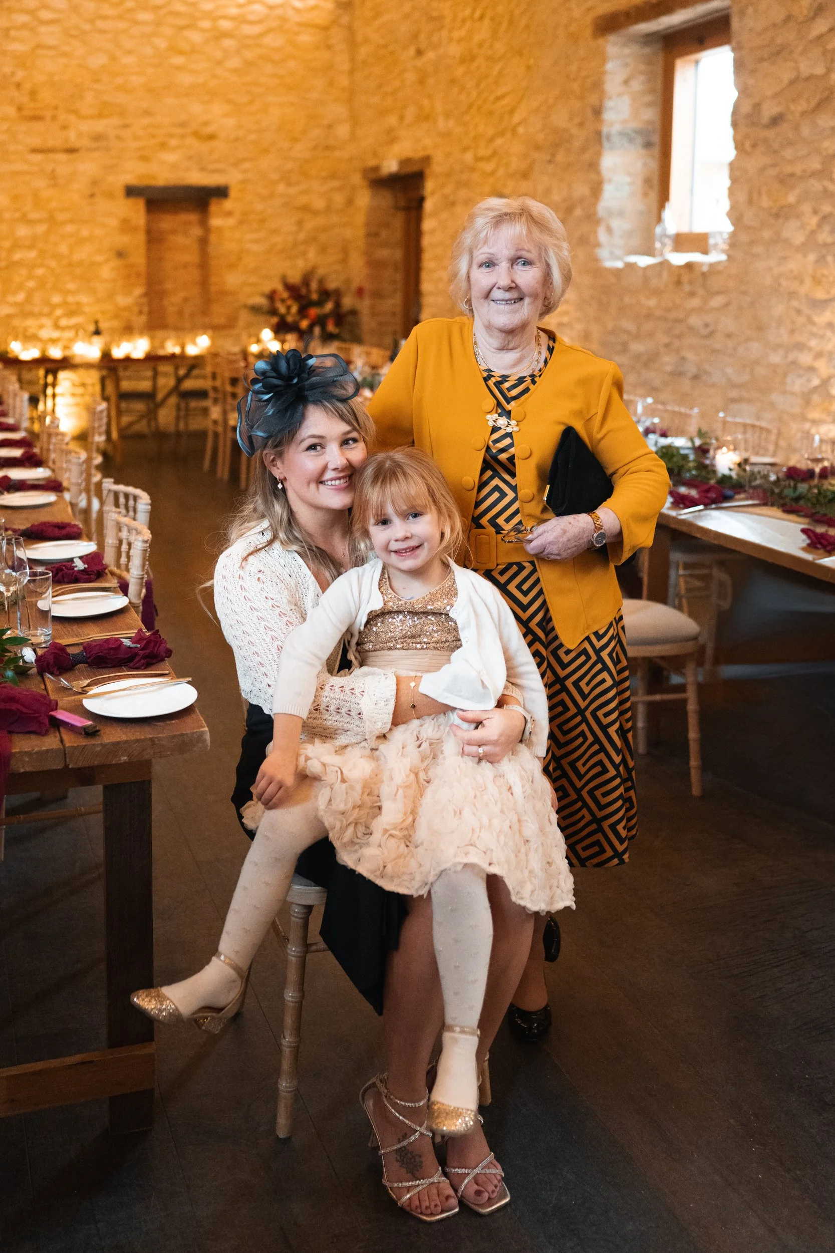 A woman, young girl, and elderly woman posing together at a decorated indoor event with dining tables in the background.