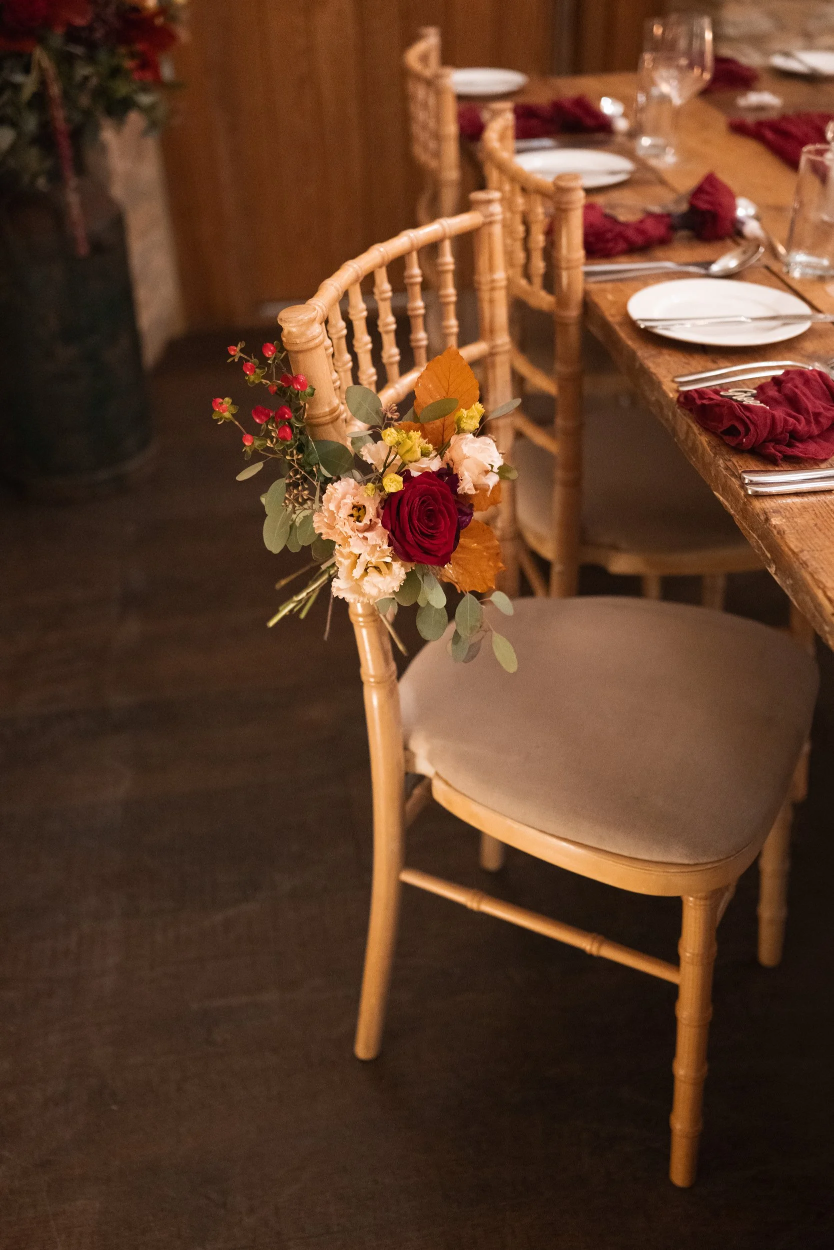 A banquet table setting with chairs, place settings, glasses, and napkins. A decorated chair has a floral arrangement with roses, fall leaves, and eucalyptus attached to its back.