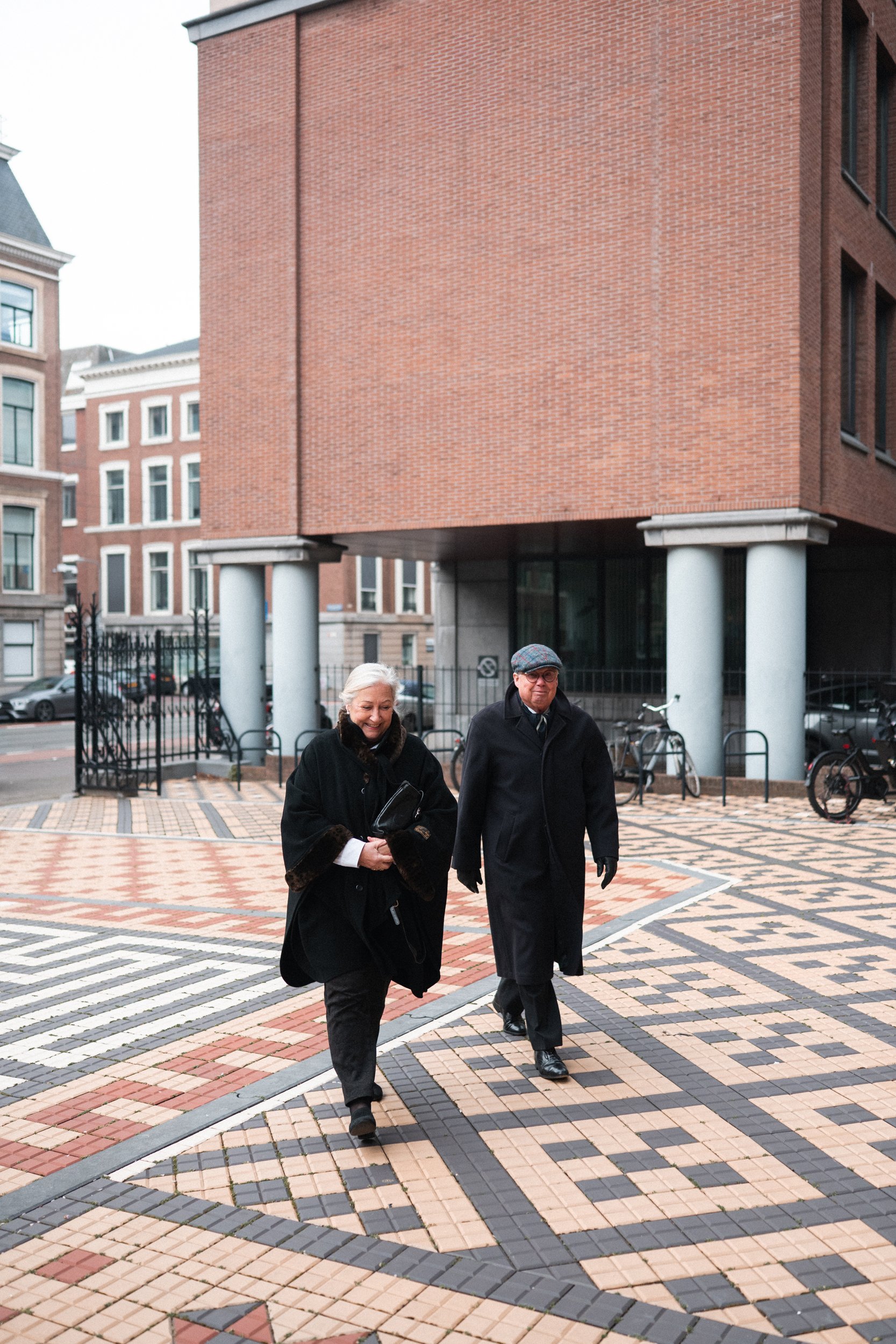 Older woman and man walking on a mosaic sidewalk in an urban area, with modern brick and concrete buildings in the background.