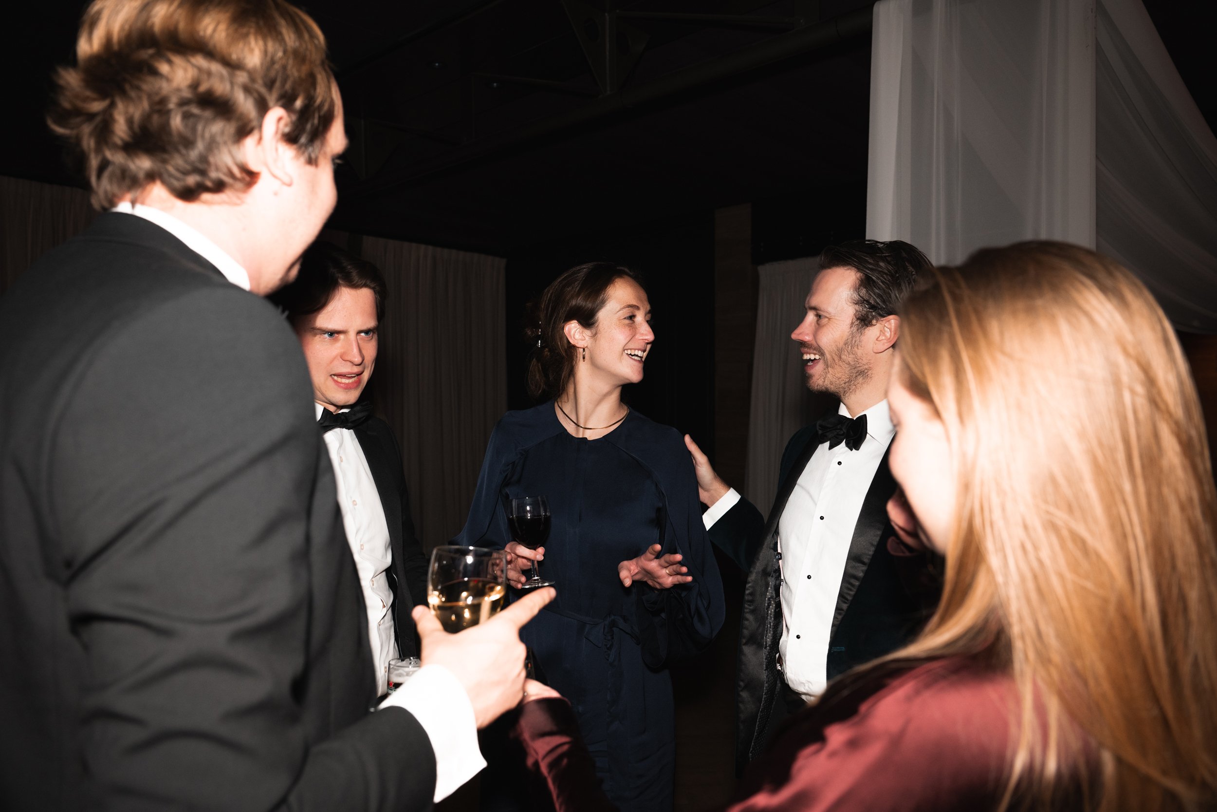 Group of five people at a formal event, four men and one woman, dressed in tuxedos and elegant attire, engaging in conversation and smiling.