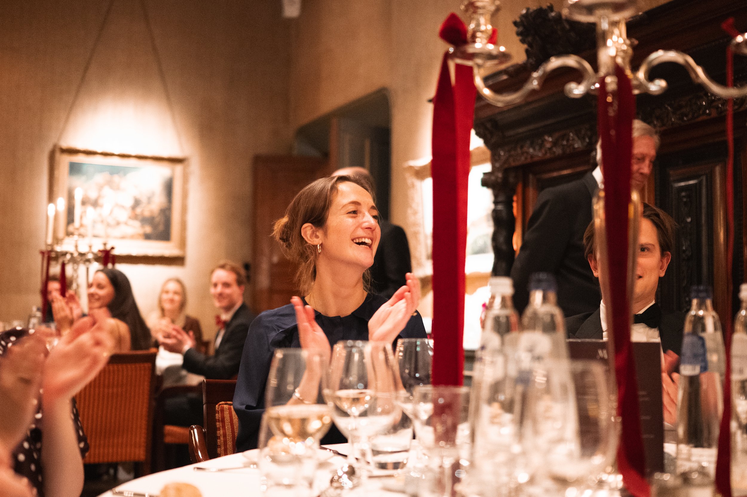 People dressed in formal attire at a dinner party, smiling and clapping, with a woman in the foreground appearing joyful. The table is set with glasses, water bottles, and decorative candles. The setting appears to be an elegant restaurant or banquet
