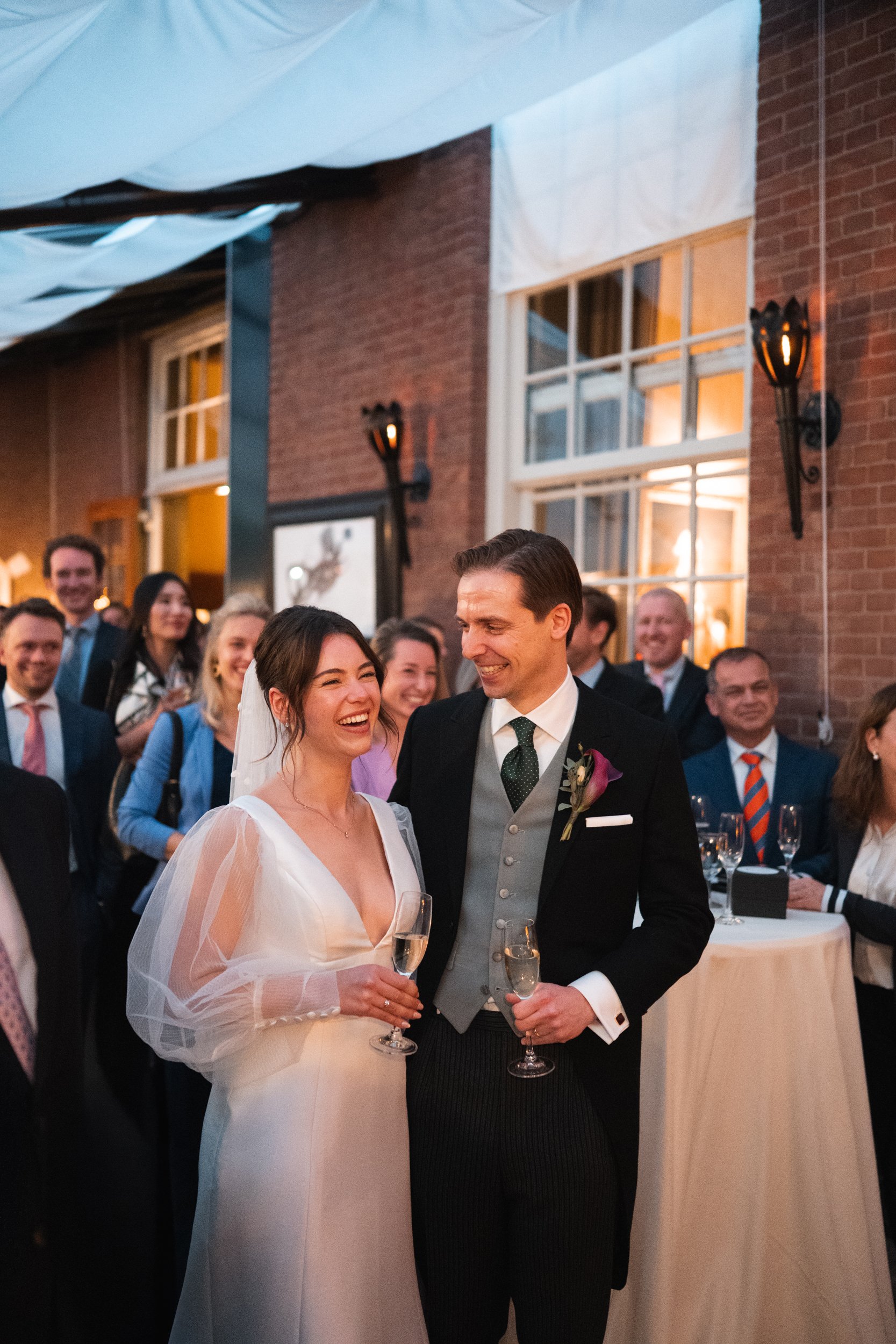 Happy bride and groom sharing a toast at their wedding reception, surrounded by friends and family in an indoor venue with brick walls and warm lighting.