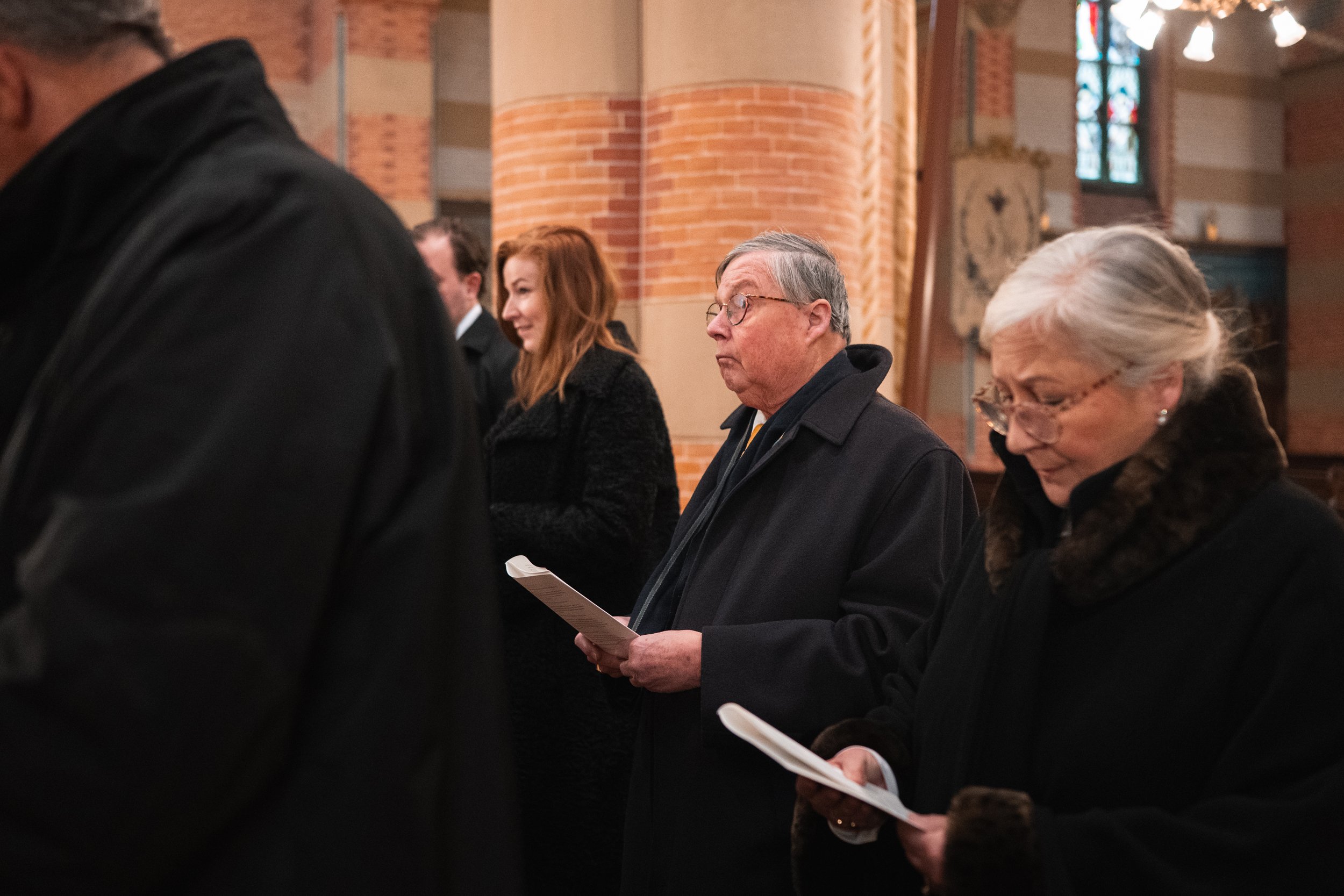 People standing in a church, holding hymn books or programs, engaged in prayer or reflection. The church has brick columns, stained glass windows, and warm lighting.