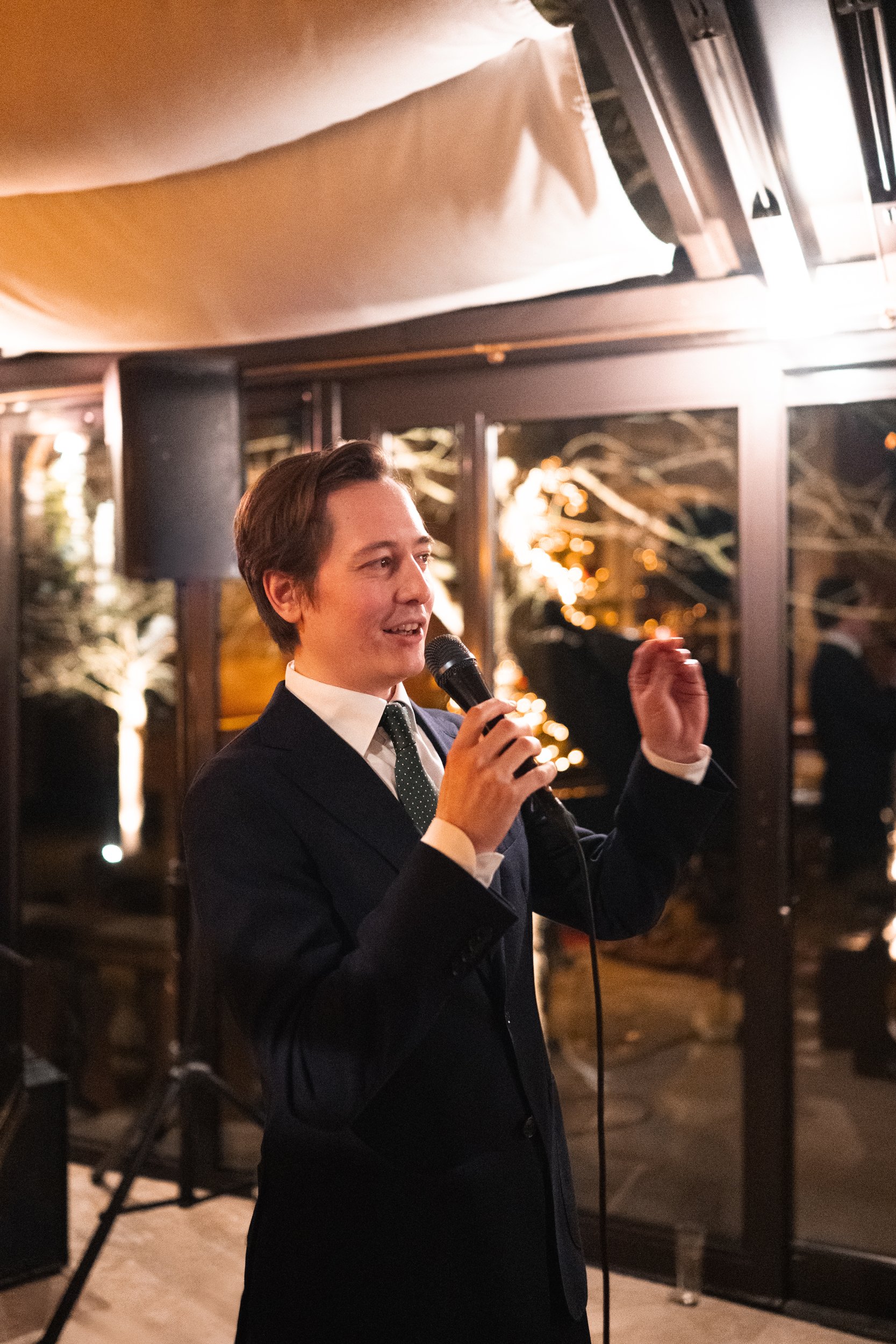 A man in a dark suit and white shirt speaking into a microphone at an indoor event, with large windows and decorative lighting in the background.