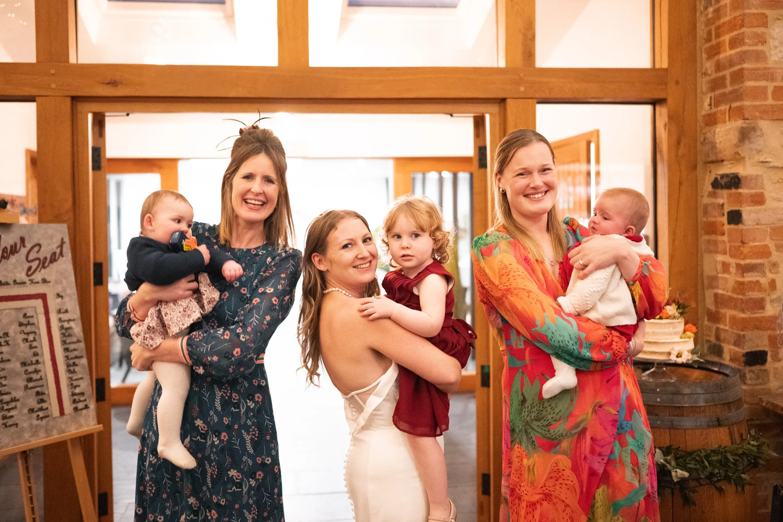Three women are smiling and holding children at a celebration indoors. The woman in the middle is wearing a white dress and holding a little girl in a red dress. The woman on the left has a blue dress and is holding a small boy, while the woman on th