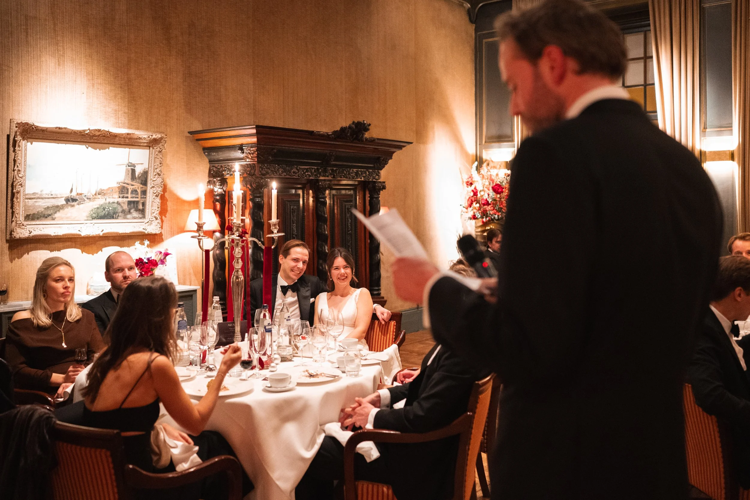 A man in a tuxedo giving a speech at a formal dinner with guests seated at a round table, including a woman in a white dress and a man in a black tuxedo, in a decorated elegant room with warm lighting, a large painting, and floral arrangements.